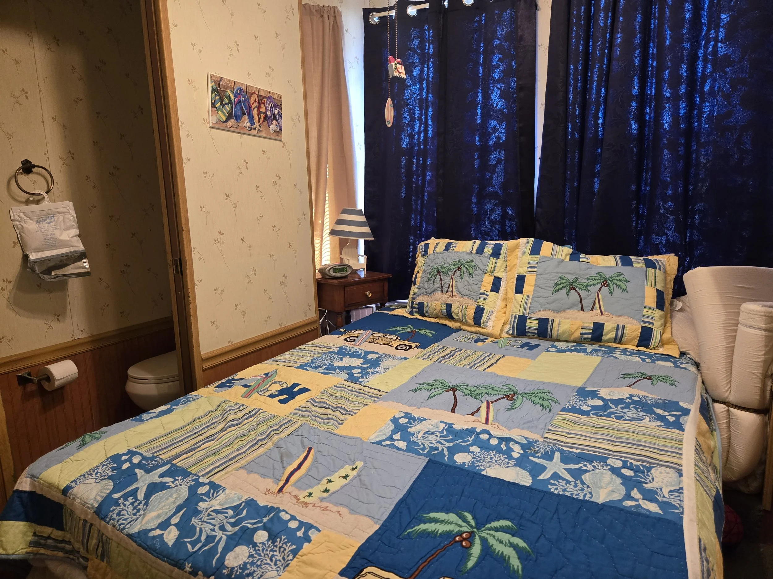 A bedroom with a bed covered in a tropical-themed quilt and pillows, dark blue curtains, a window with beige curtains, a nightstand with a striped lamp, and a wall with colorful artwork.