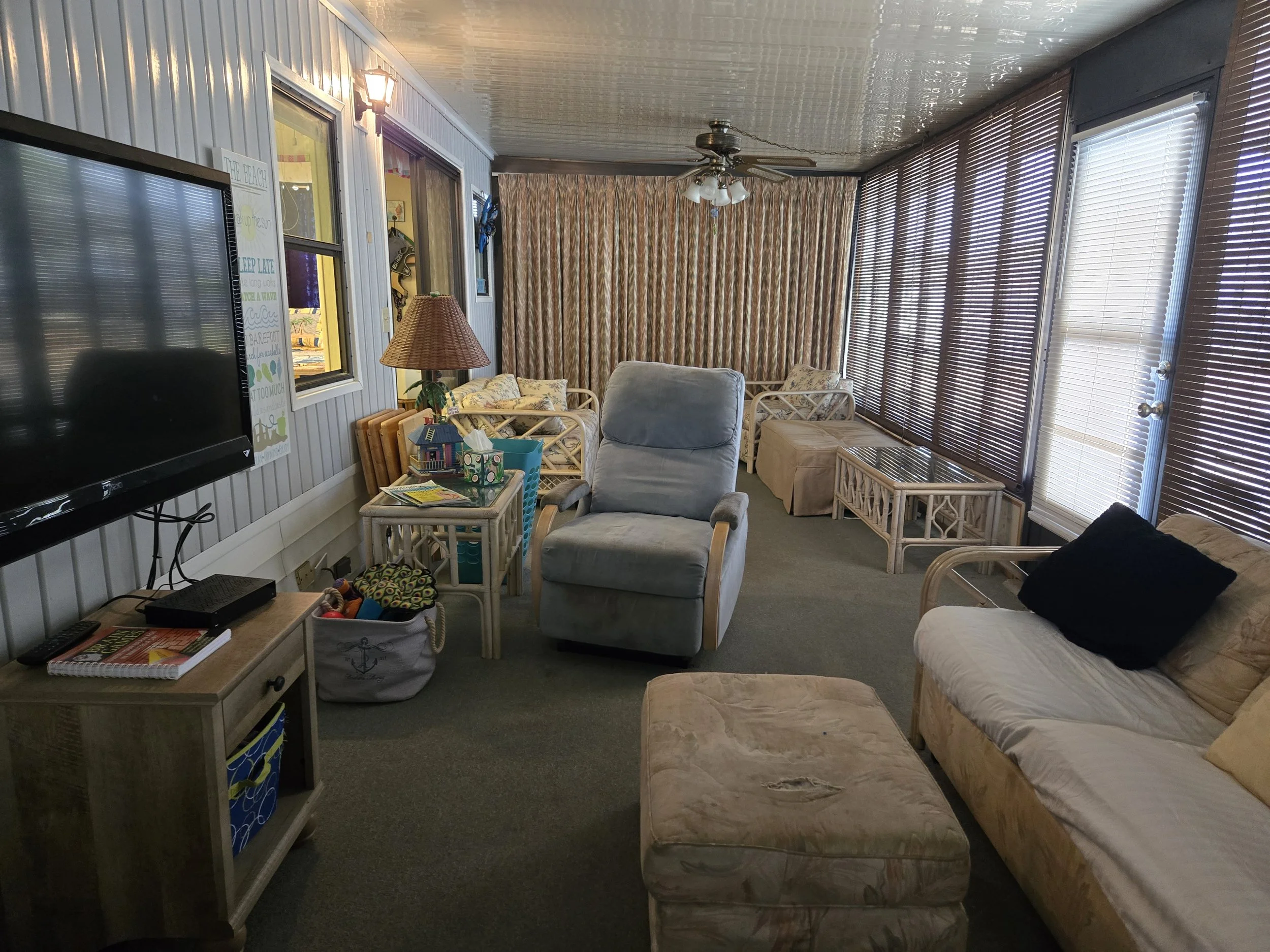Living room with beige and tan furniture, a wall-mounted TV, a table with toys, a window with brown blinds, and a ceiling fan.