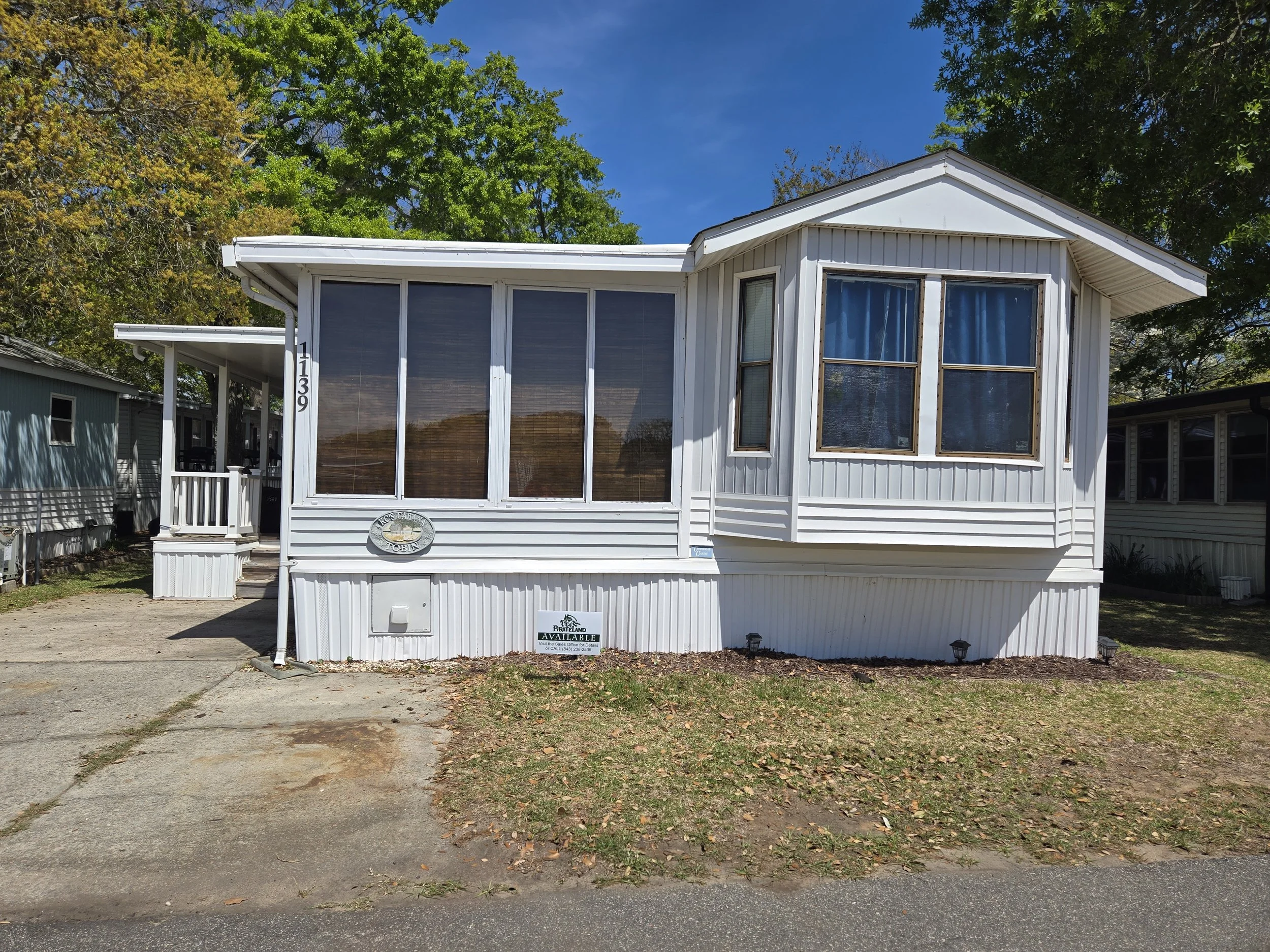 A white manufactured home with a screened porch and large windows, daytime with clear blue sky and surrounding trees.