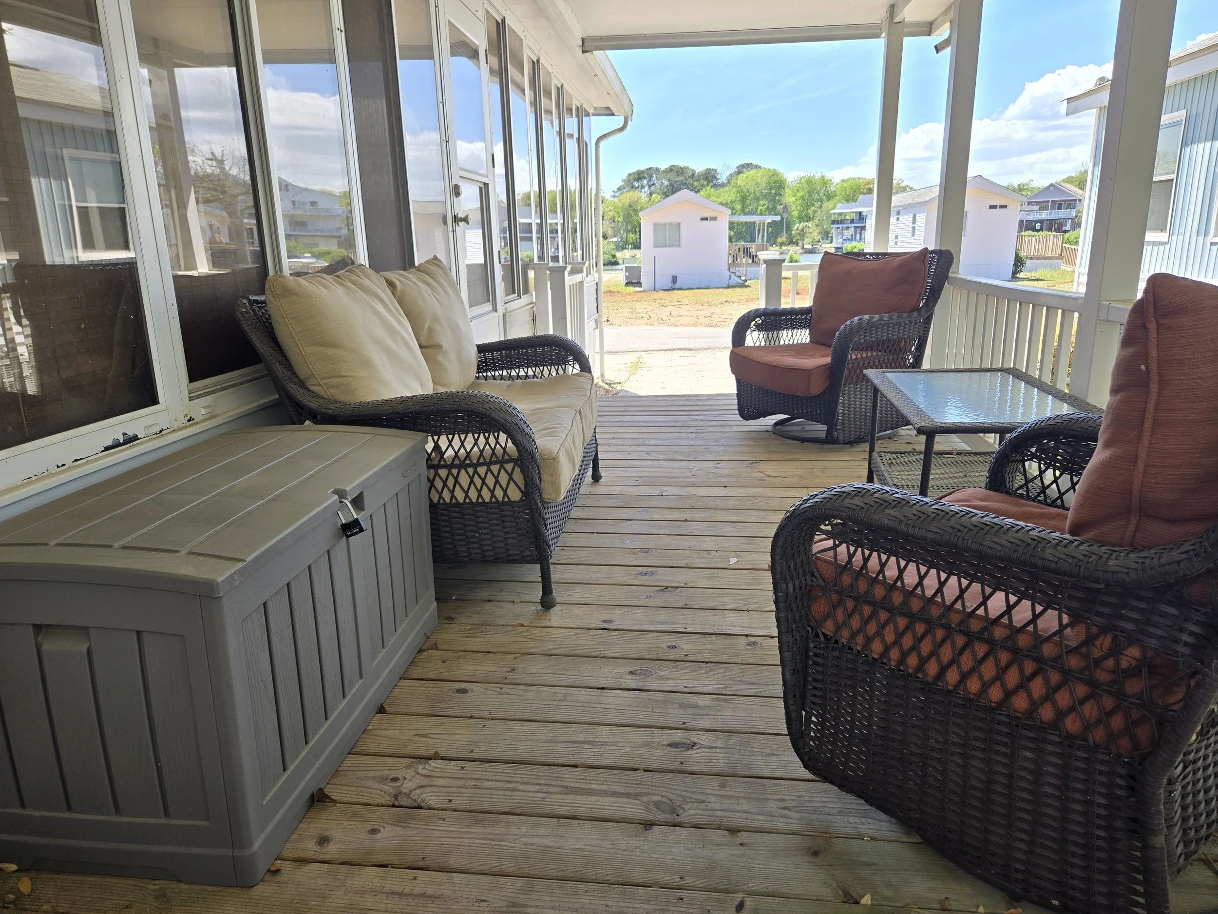 Porch with wicker seating, cushions, a storage chest, and outdoor tables, overlooking neighboring houses and a yard on a sunny day.