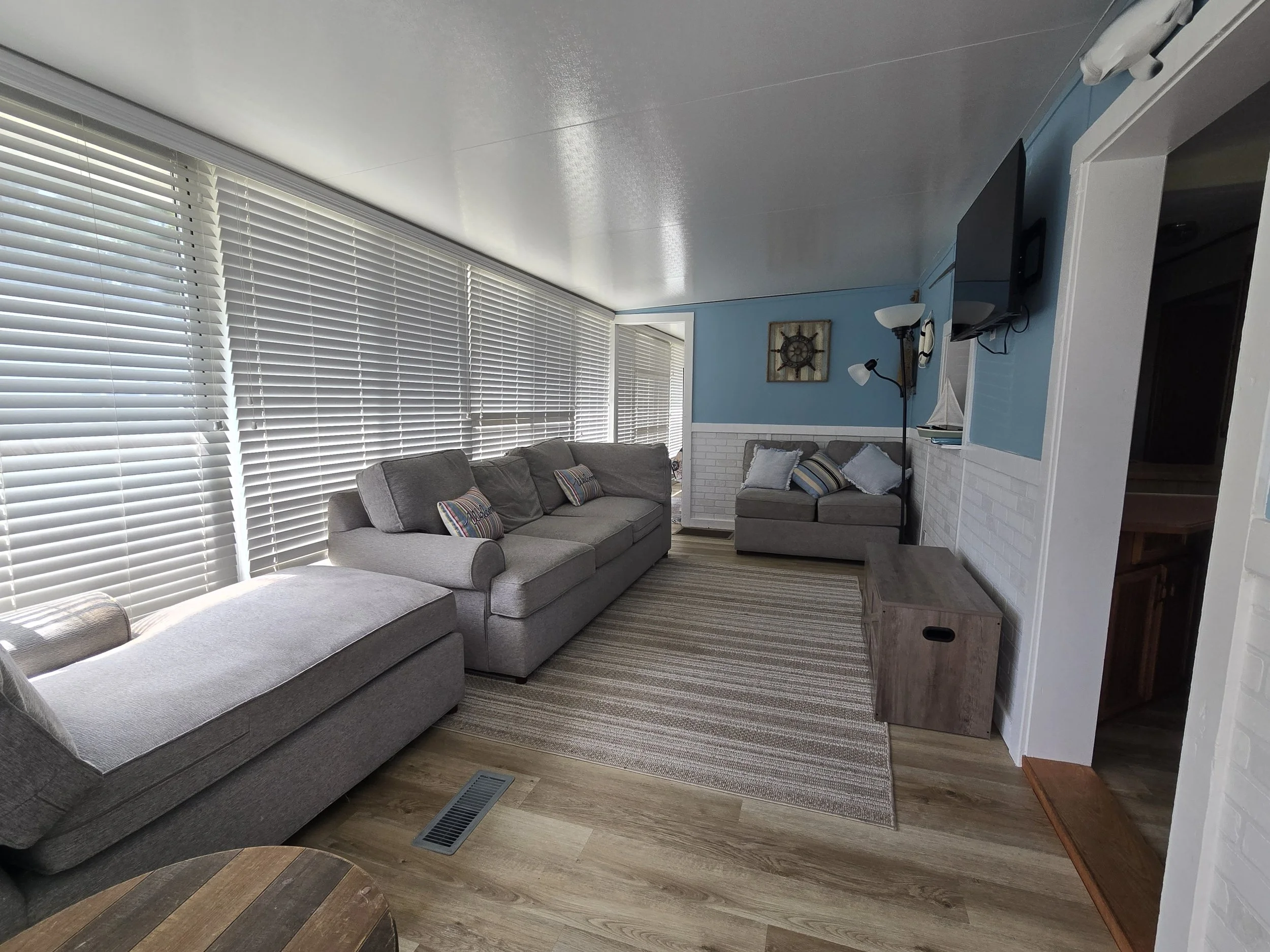 Living room with large windows covered by white blinds, gray sectional sofa with striped pillows, a striped area rug, a wooden side table, a TV mounted on a blue wall with nautical decor, and a white brick accent wall.