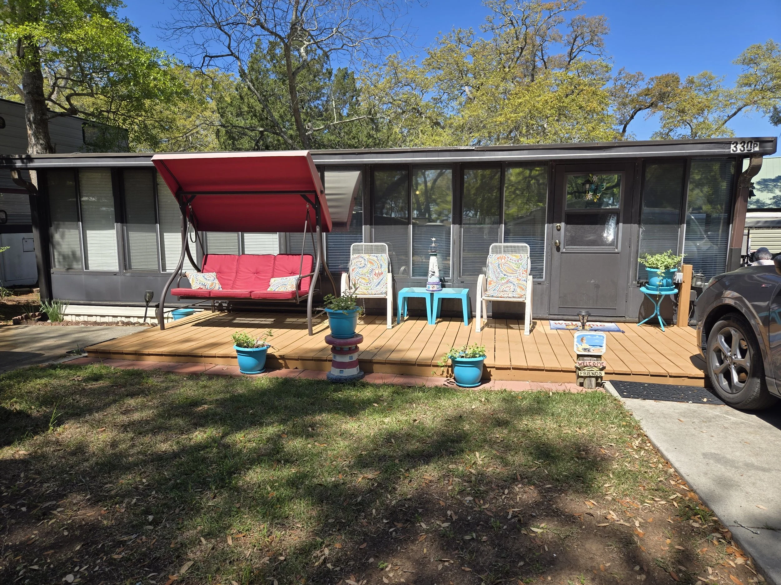 Backyard porch with a black mobile home, featuring a red porch swing with two pillows, two white patio chairs, a small blue table with a lighthouse decor, and potted plants in blue containers. There is a sign with welcoming messages, and a car is par