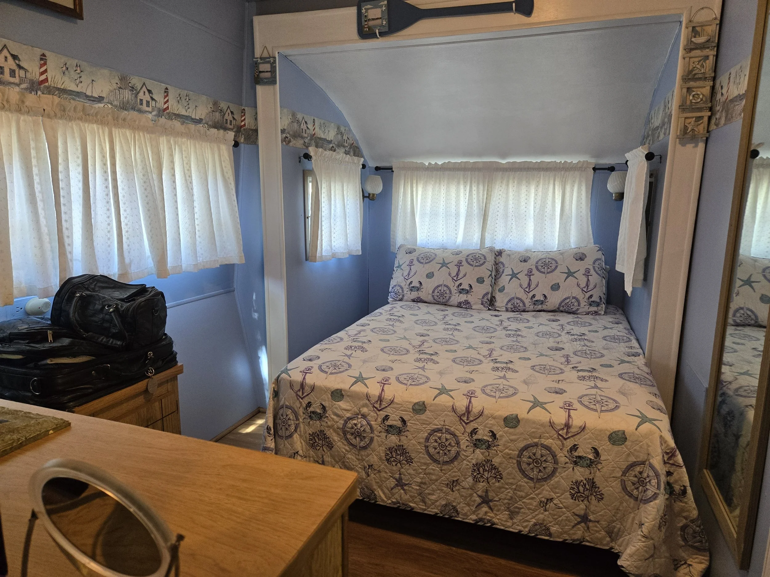 A cozy bedroom with blue walls, with a bed covered in a nautical-themed quilt and pillows, two windows with white curtains, and a mirror on the right wall.