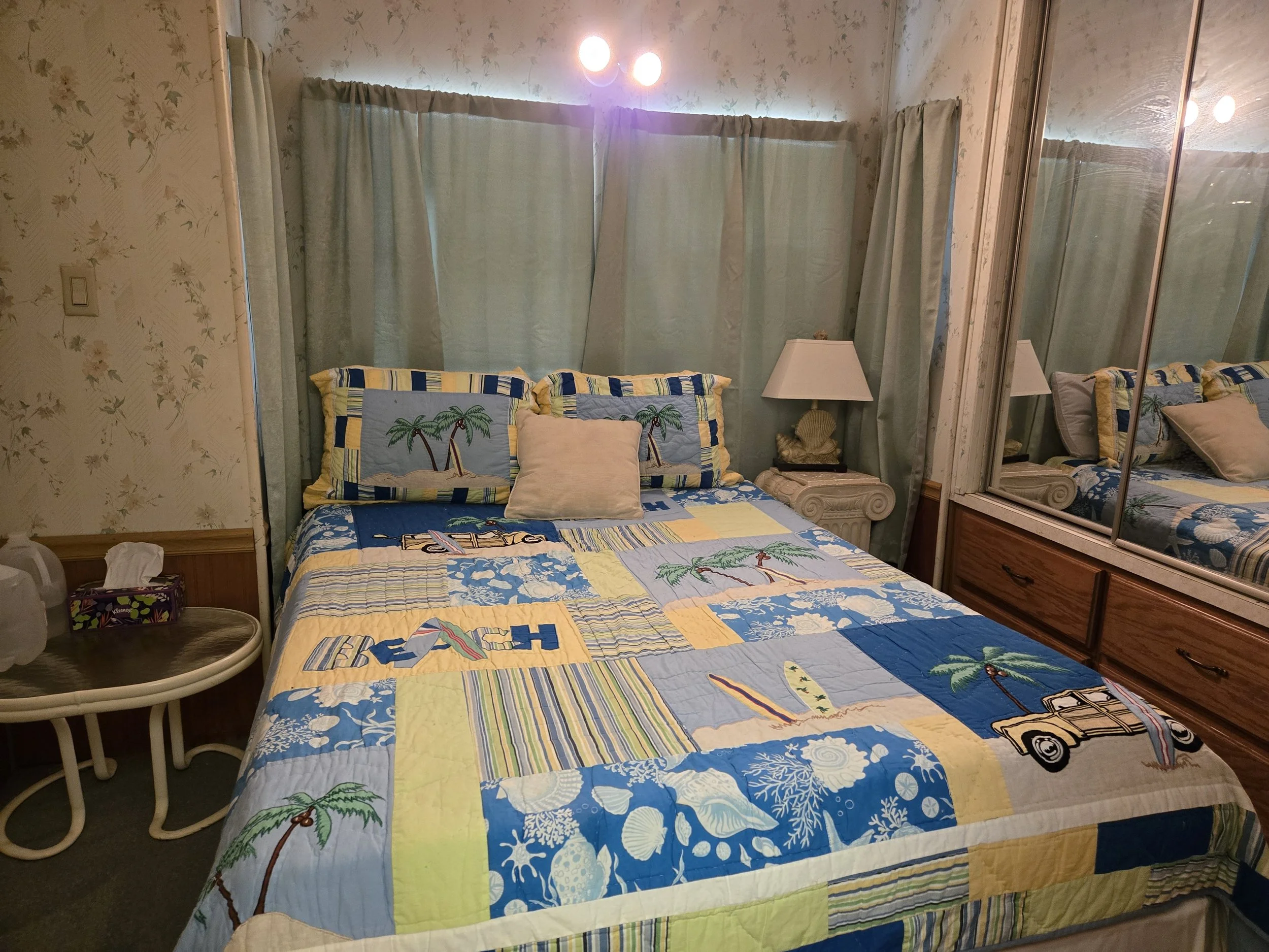 A bedroom with a bed that has a Marine-themed quilt and pillowcases, light green curtains, a mirrored closet, a white lamp on a small table, a tissue box on an end table, and reflected light from ceiling fixtures.