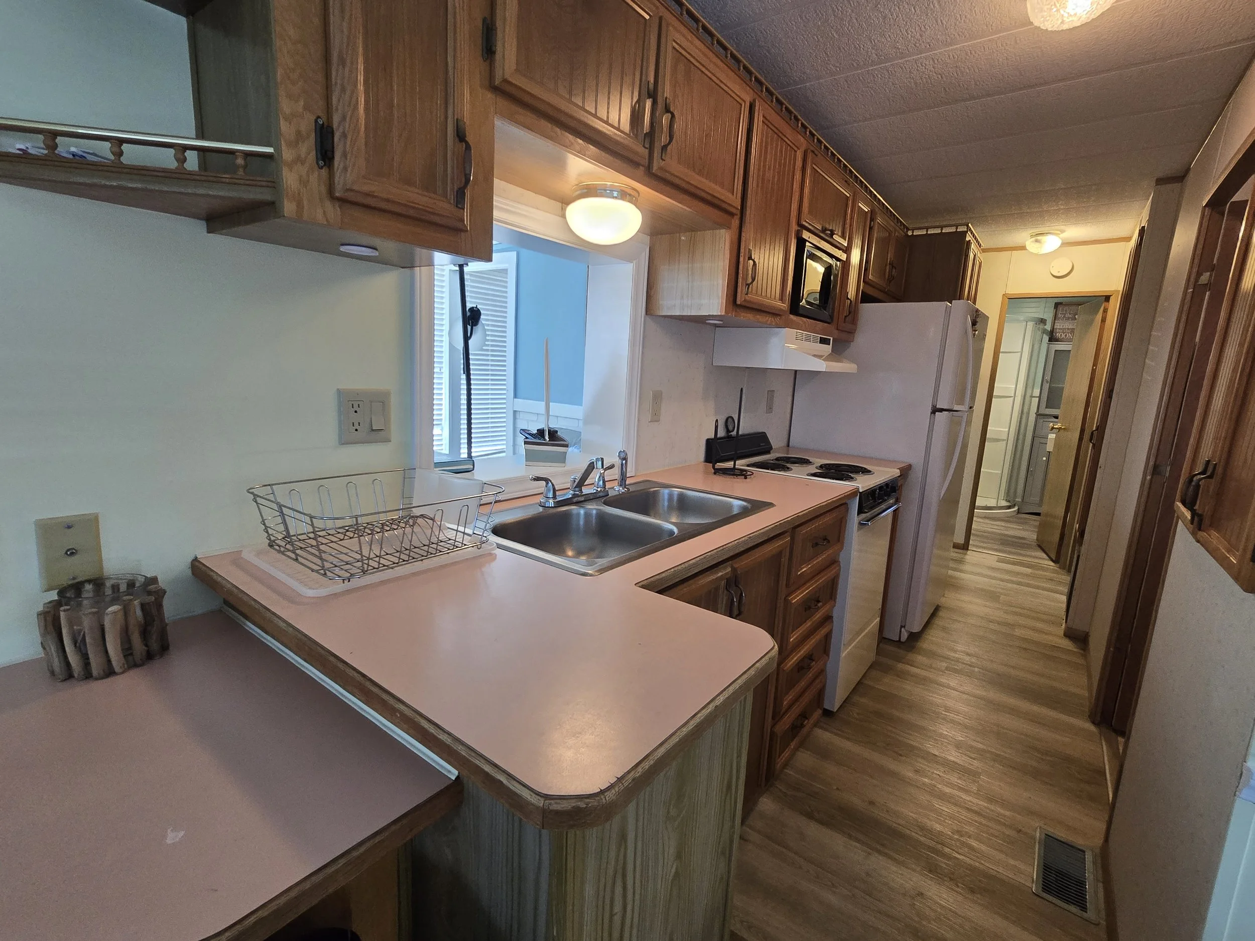 A kitchen with wooden cabinets, a pink countertop, a double sink, a stovetop, and a refrigerator. There is a window with blinds and a ceiling light fixture.