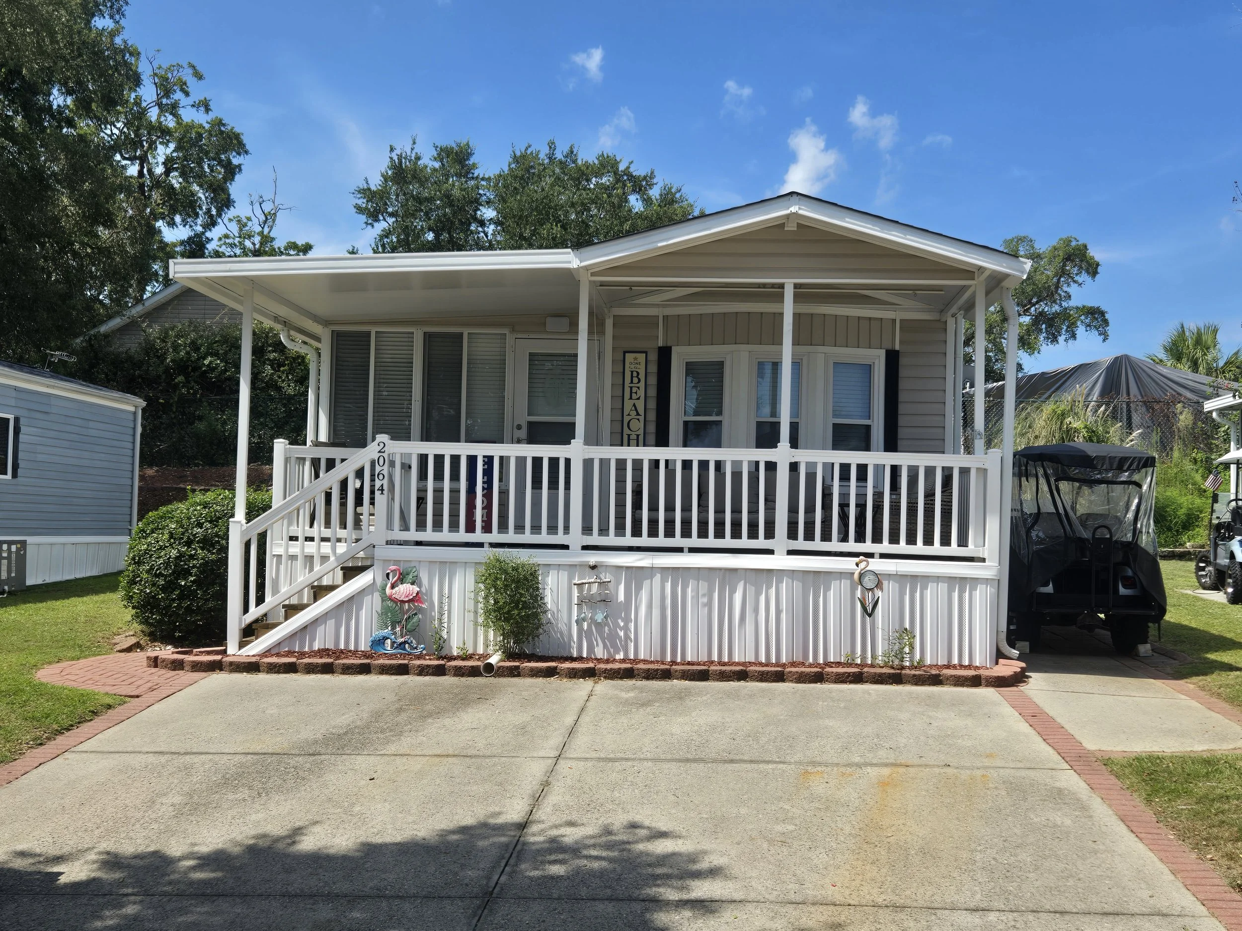Front view of a house with a porch, white railing, and a sign that says 'Beach' hanging next to the door. There's a small garden area with decorative flamingos and a paved walkway leading to the steps. A golf cart is parked on the driveway to the rig