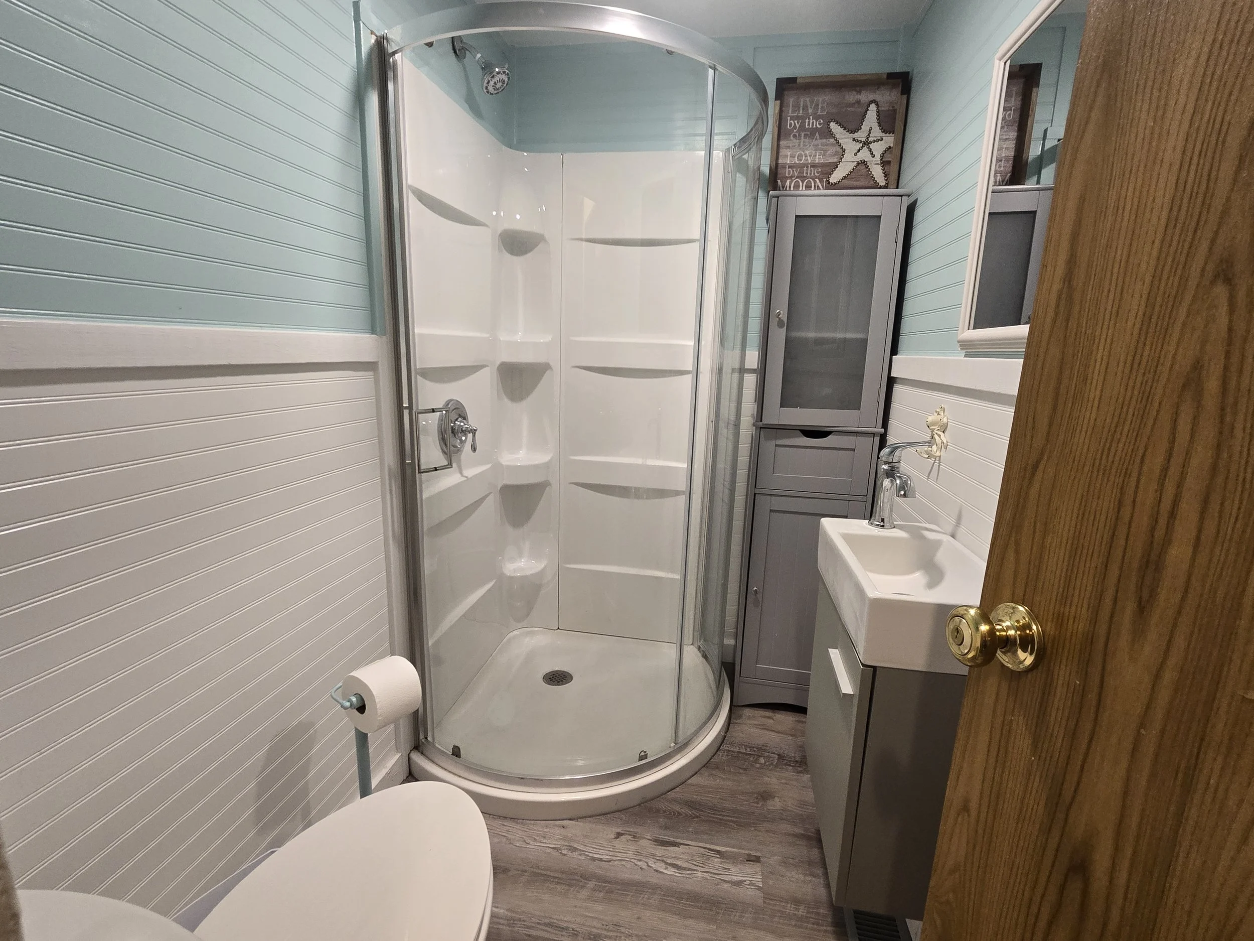 A small bathroom with a corner shower with curved glass doors, a gray cabinet, a small sink, a mirror, and a wall decoration with starfish and reads, "Live by the sea, love by the moon."