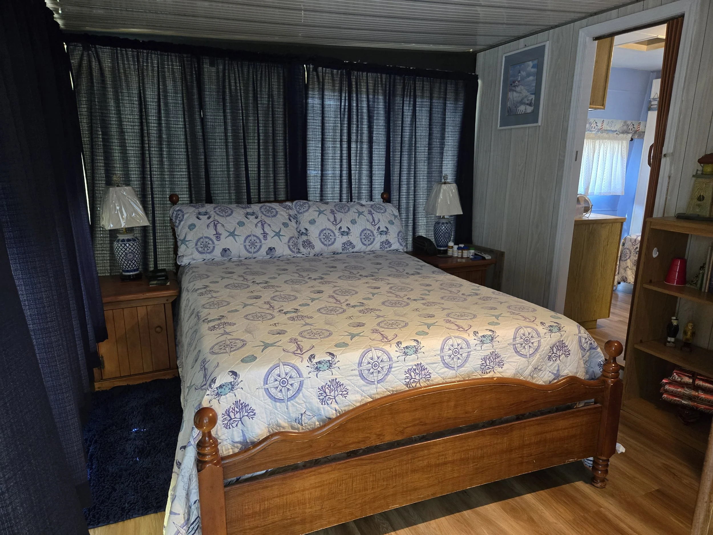 A bedroom with a wooden bed, nautical themed quilt and pillows, side tables with lamps, and window with black curtains.