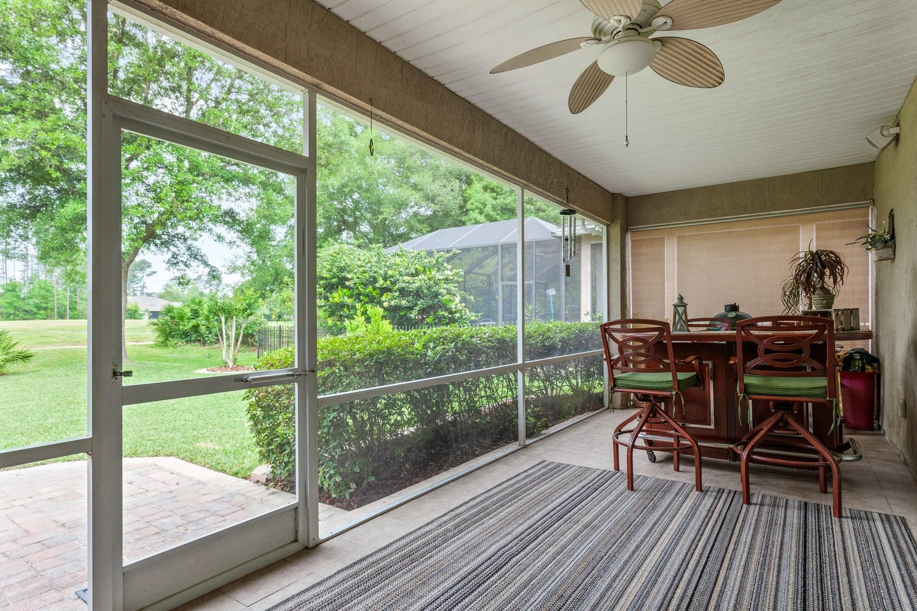 The interior of a screened-in porch with a striped rug and bar with two barstools inside on the right and a yard with bushes and trees outside of the screen wall and door on the left.