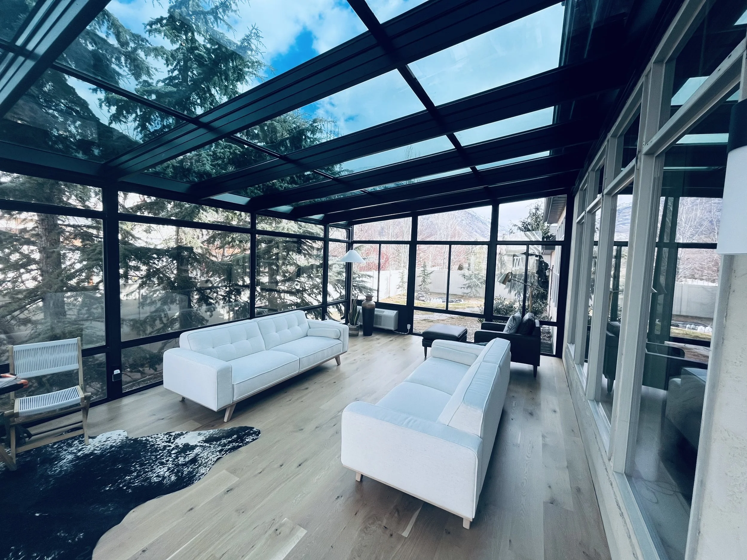 Sunroom with large glass windows, white couches, black chairs, a cowhide rug, and indoor plants, overlooking a winter landscape with trees and mountains.