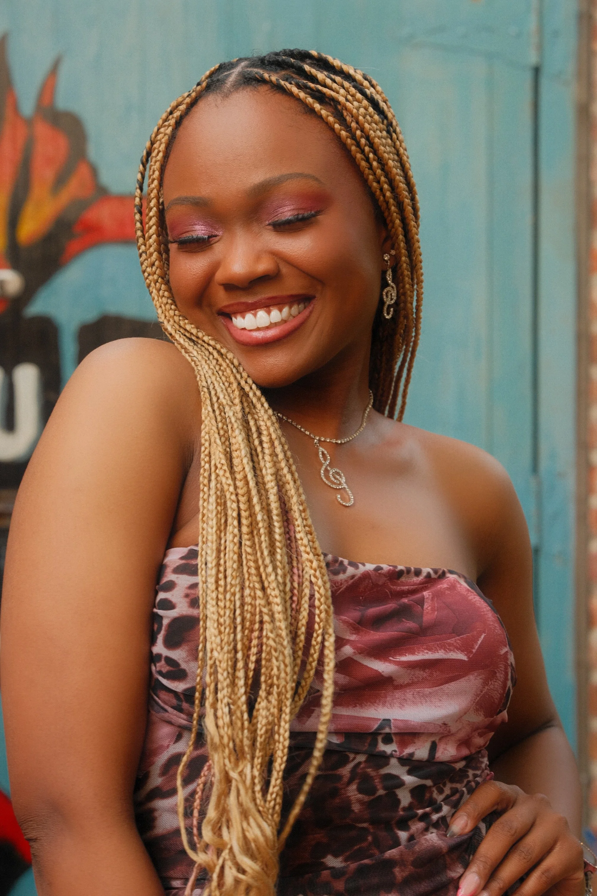 Chloé V smiling in with her hand on her hip in front of the House of Blues logo in New Orleans while wearing a pink and brown floral strapless dress. She has long blonde braids that are laying over her right shoulder (on the left from the photographe