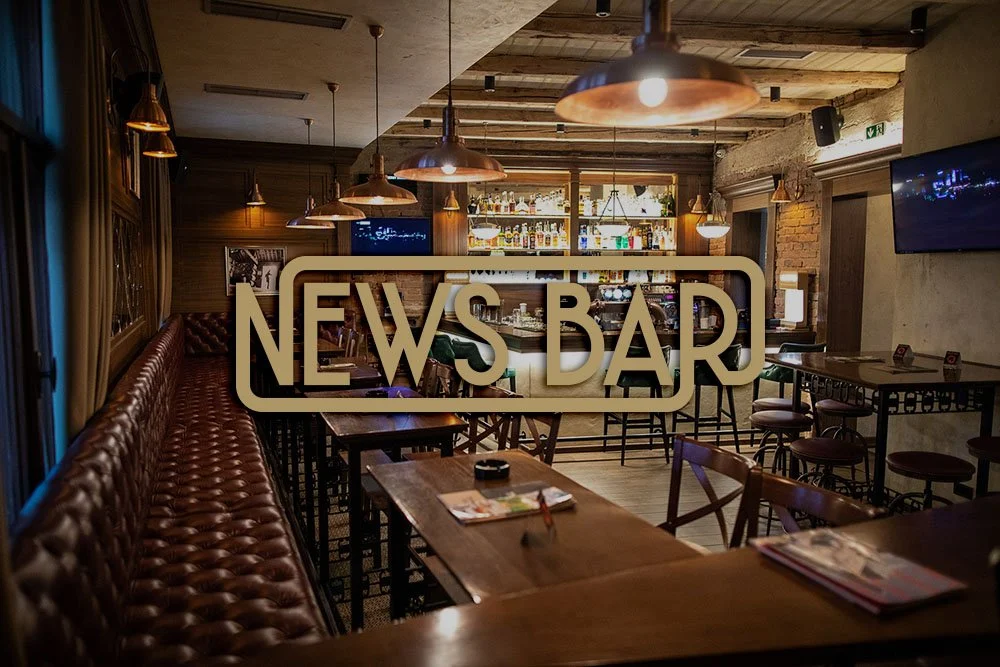 Interior of a cozy pub or bar with wooden tables, bar counter with bottles, leather seating, pendant lighting, and a television mounted on the wall.
