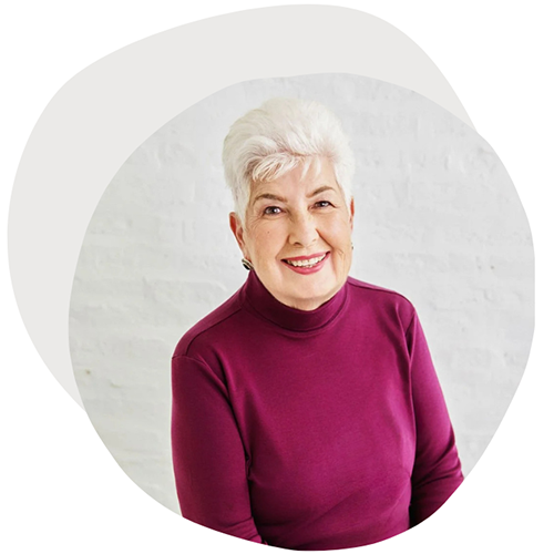 An older woman with short white hair, smiling, wearing a purple turtleneck, standing in front of a white brick wall.
