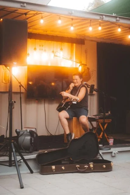A singer and guitarist performing beneath warm lights at an outdoor live-music event