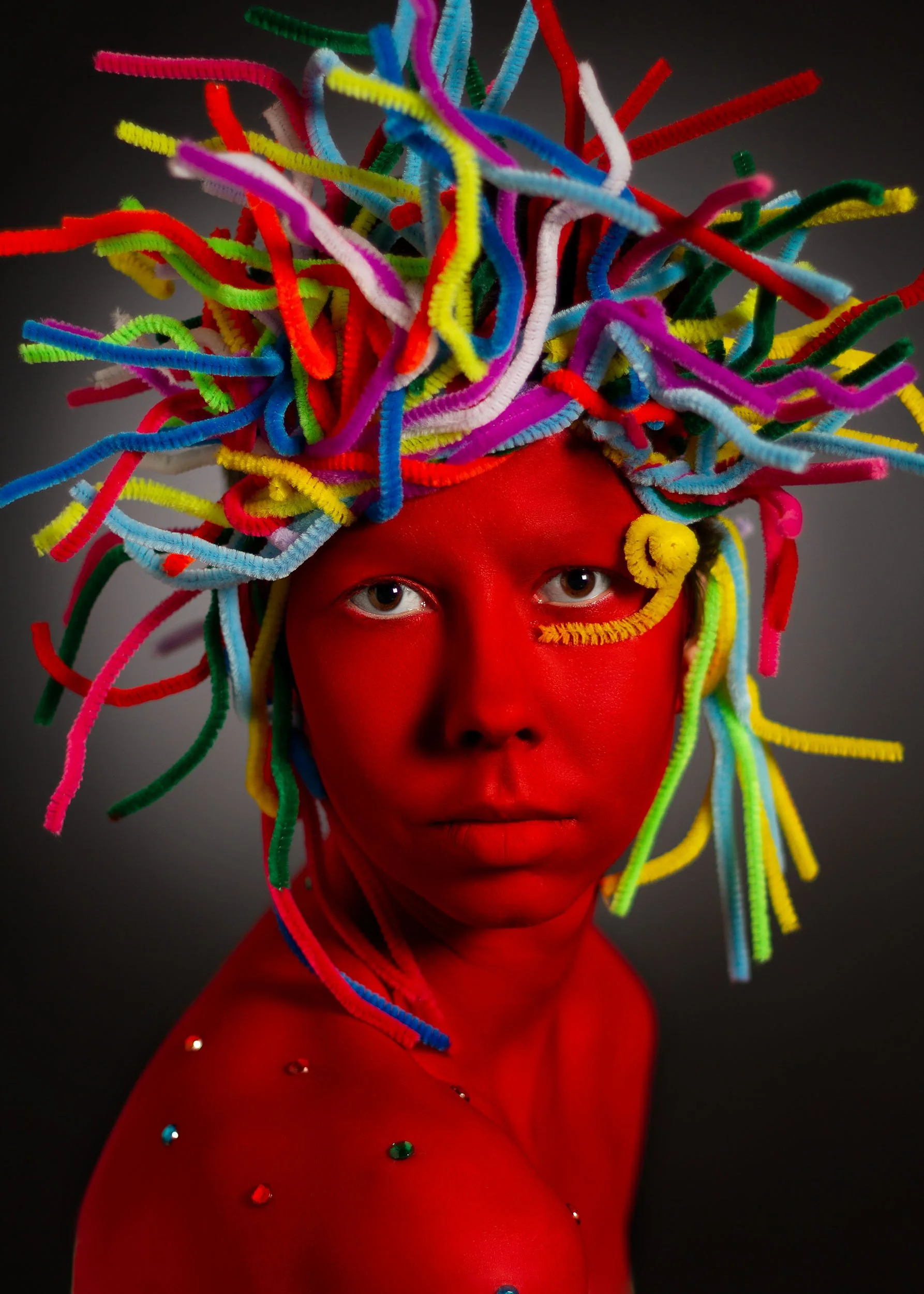Une personne avec la peau entièrement peinte en rouge et des cheveux faits de pipe cleaners multicolores enchevêtrés, se tenant contre un fond sombre.