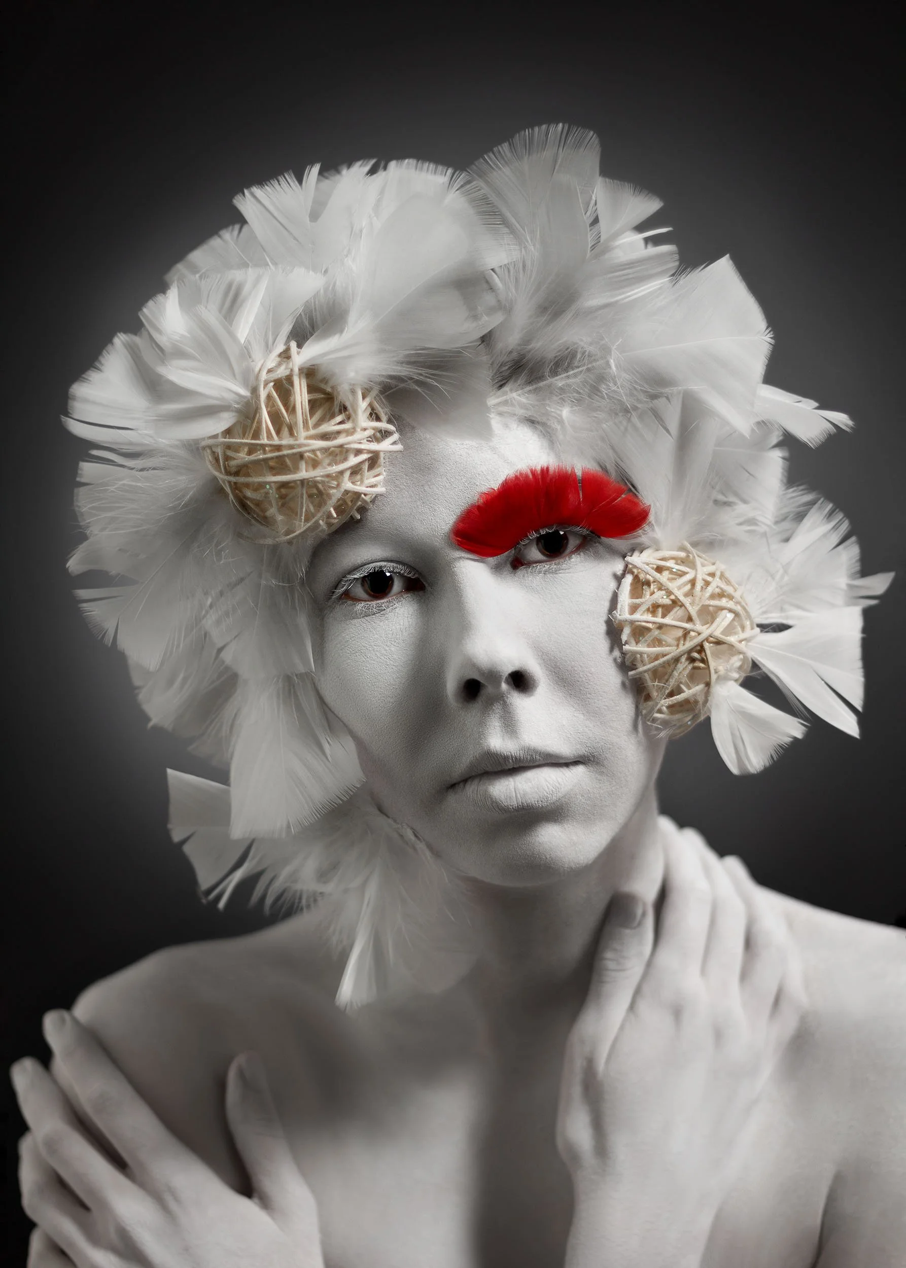Portrait d'une femme avec un maquillage dramatique, portant ornement de plumes blanches et de décorations tressées, avec une plume rouge autour de l'œil.