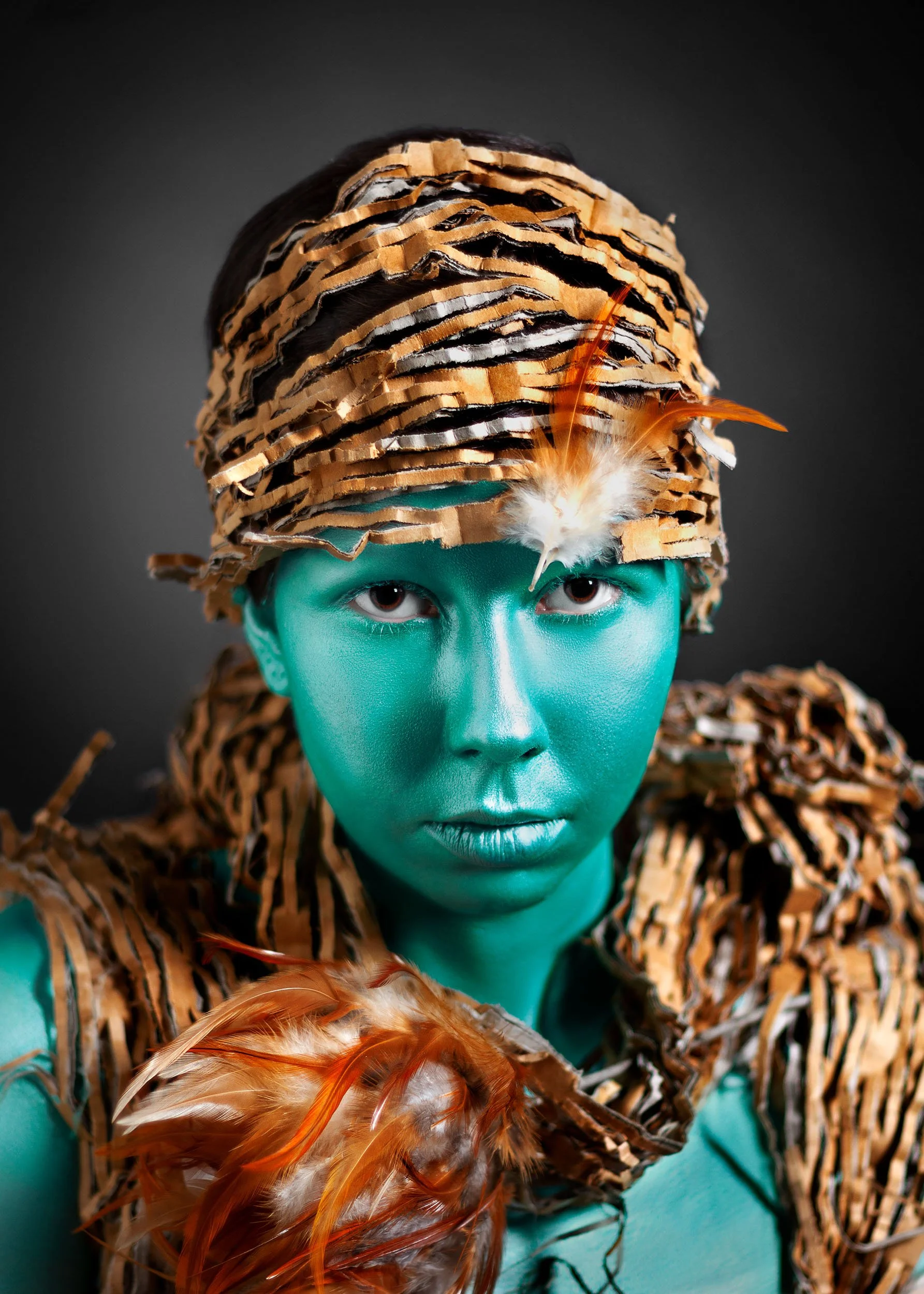 Portrait d'une personne avec la peau turquoise, portant un costume et un couvre-chef faits de plumes et de matériel recyclés, avec un fond sombre.