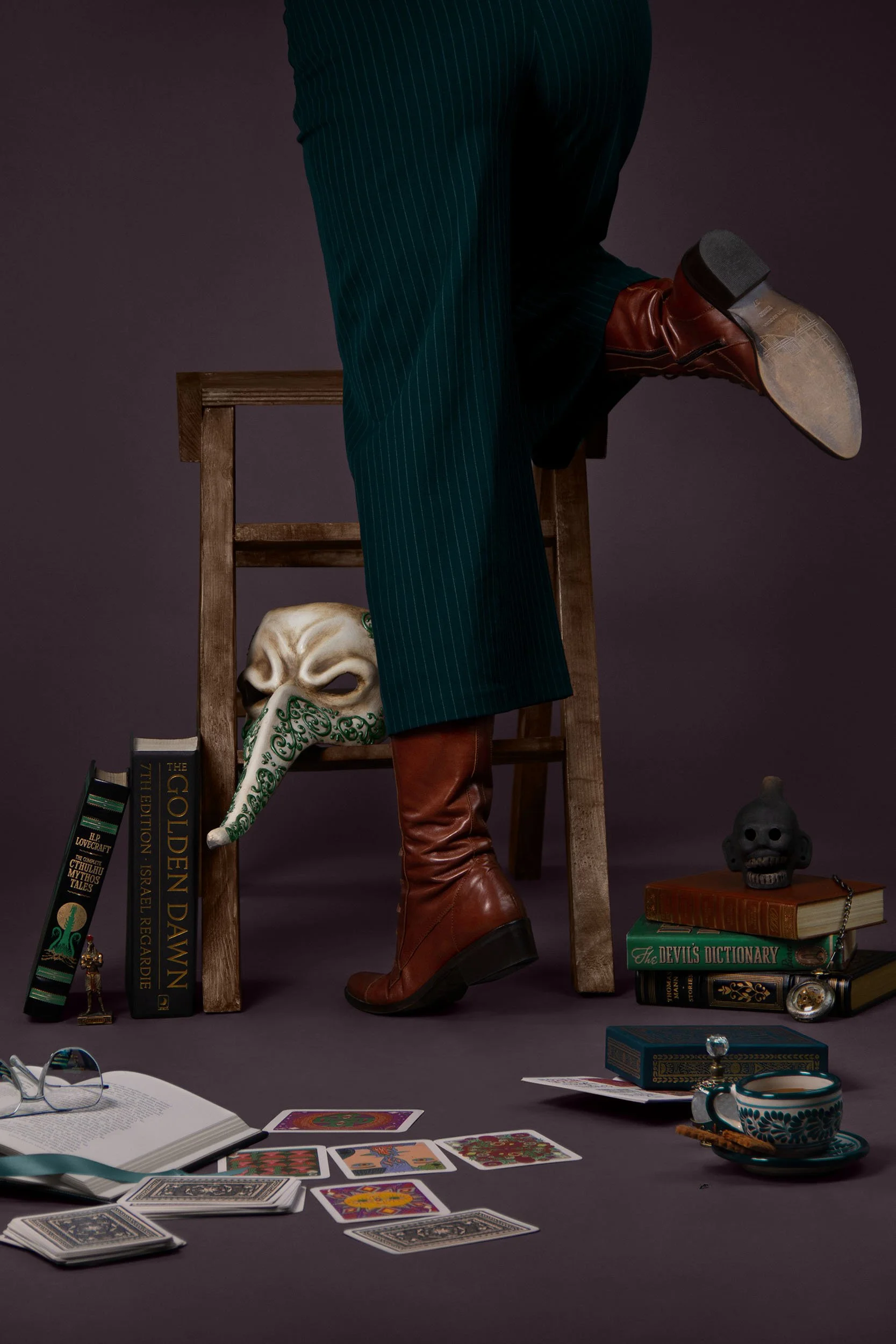 Une scène avec une personne montant sur un escabeau, portant des bottes en cuir marron, un pantalon à rayures. Autour d'elle, il y a des livres, des cartes de tarot, une tasse à thé, des lunettes et d'autres objets mystiques.