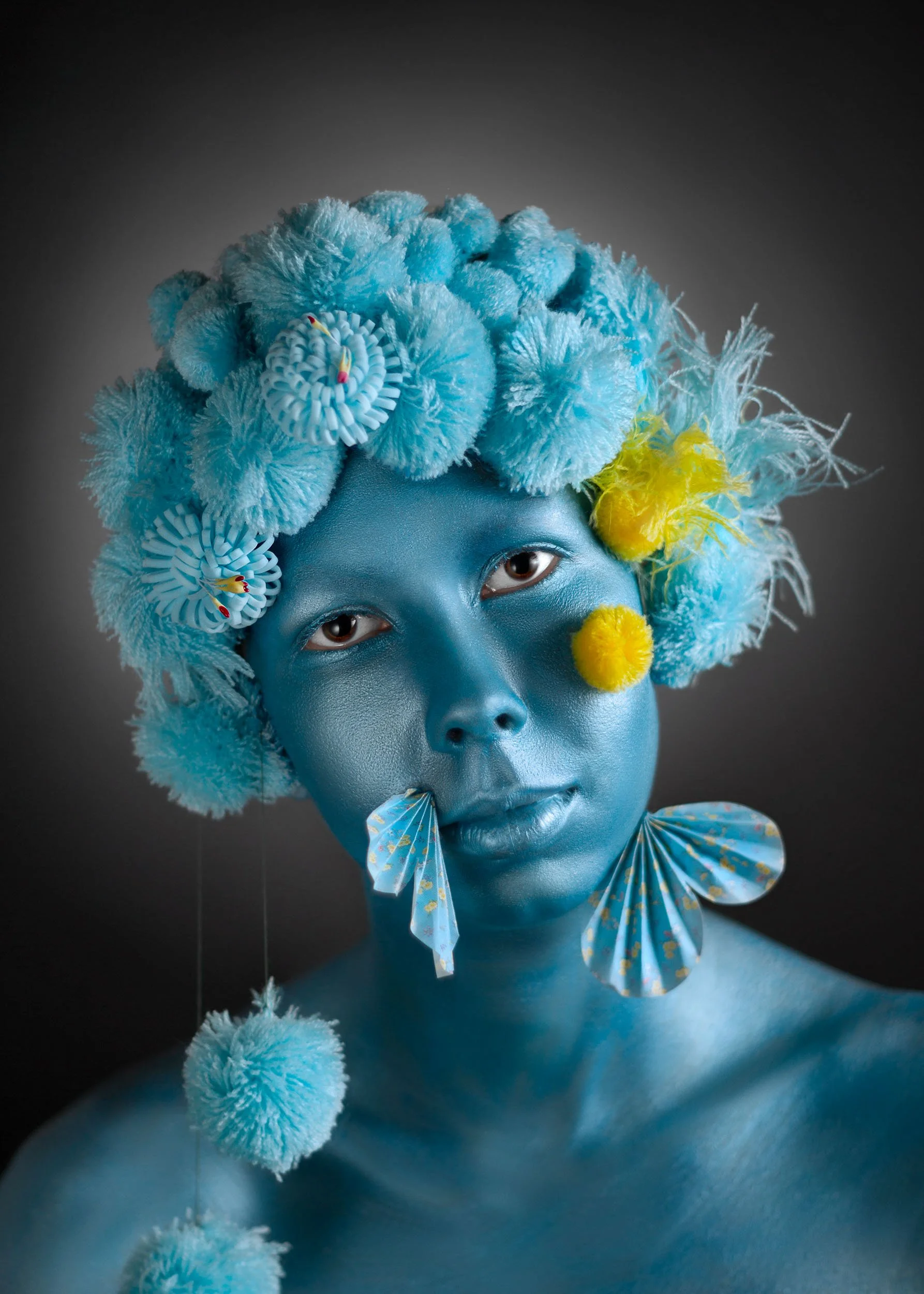 Portrait d'une personne au visage peint en bleu, portant des accessoires en forme de papillons en papier bleu avec des motifs jaunes, des pompons jaunes et des décorations en forme de fleurs, avec un fond noir.