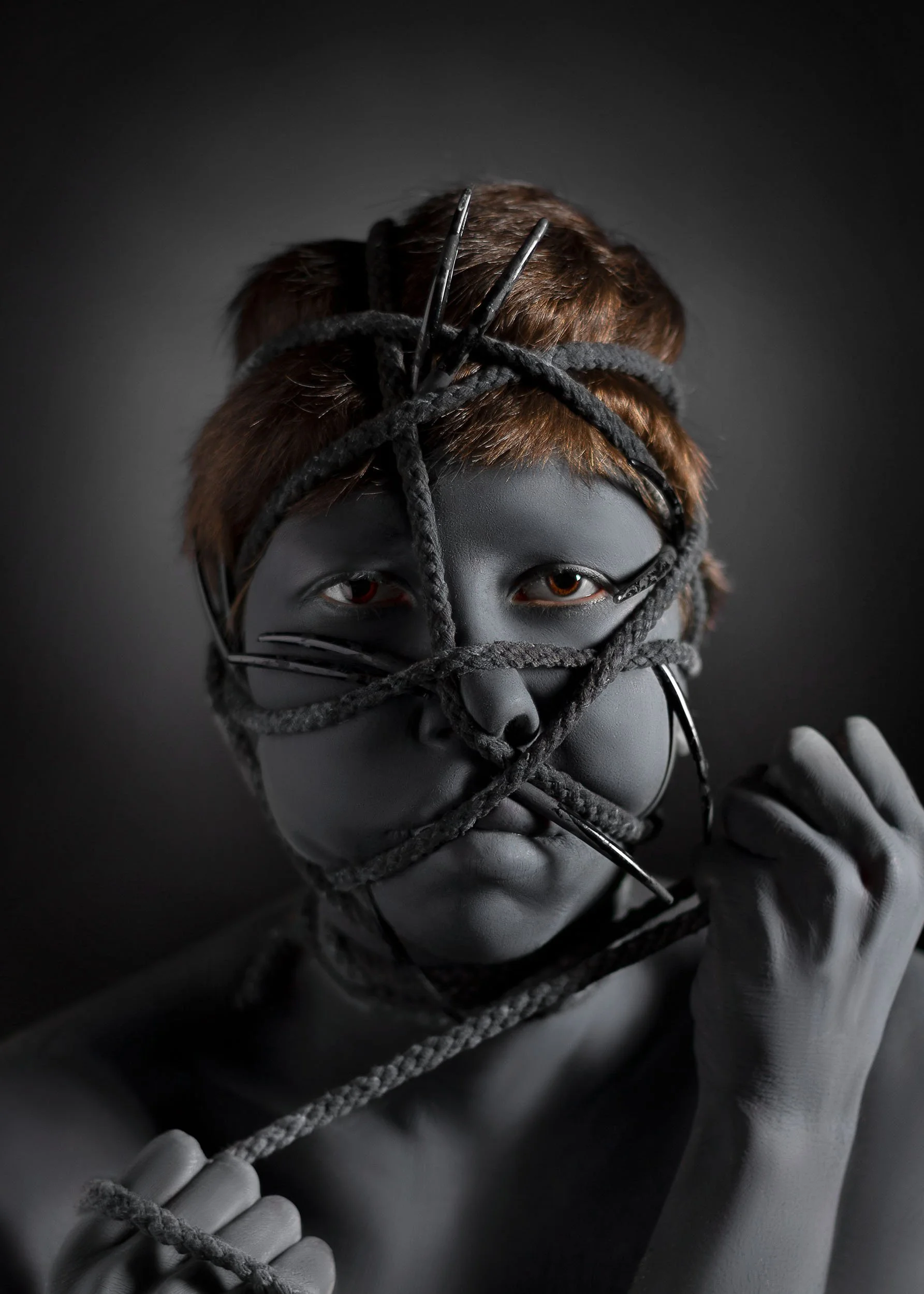 Une personne avec la peau entièrement peinte en noire, une corde noirs et gris enroulés autour de sa tête et de son visage, avec un fond noir.