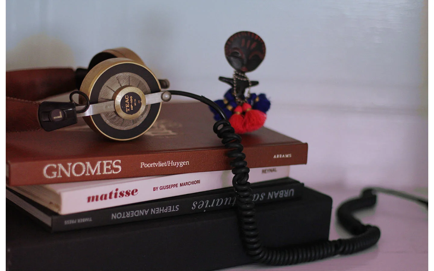 Close-up of a stack of books with a vintage headset and a small colorful doll on top. The books are titled "GNOMES" by Poortvliet/Huygen, "matisse" by Giuseppe Marchiori, and "Steven Anderson." The background is plain white.