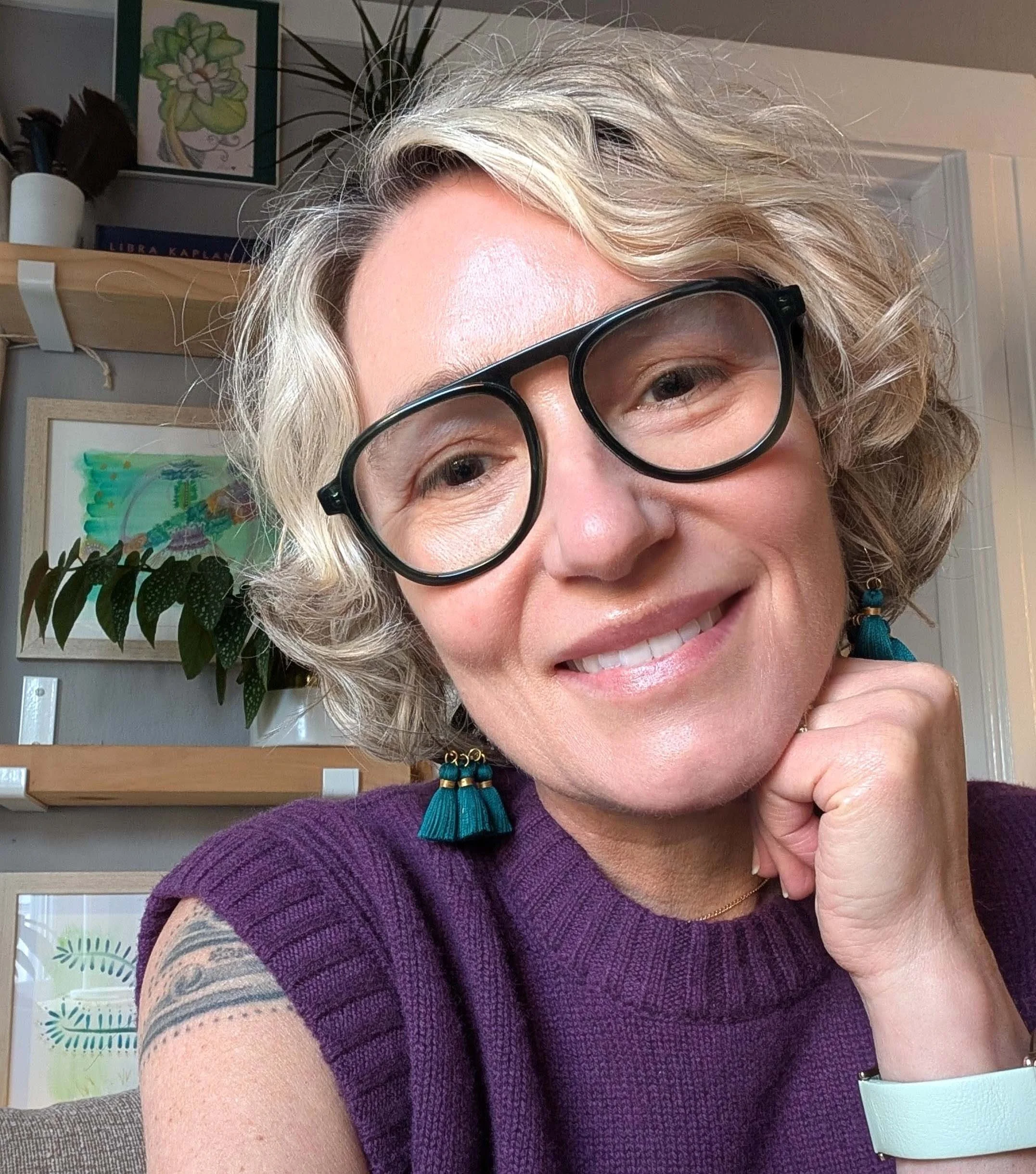 Close-up of a smiling woman with curly blond hair, wearing large black glasses, turquoise earrings, and a purple textured top. She is indoors, with artwork, plants, and a shelf in the background.