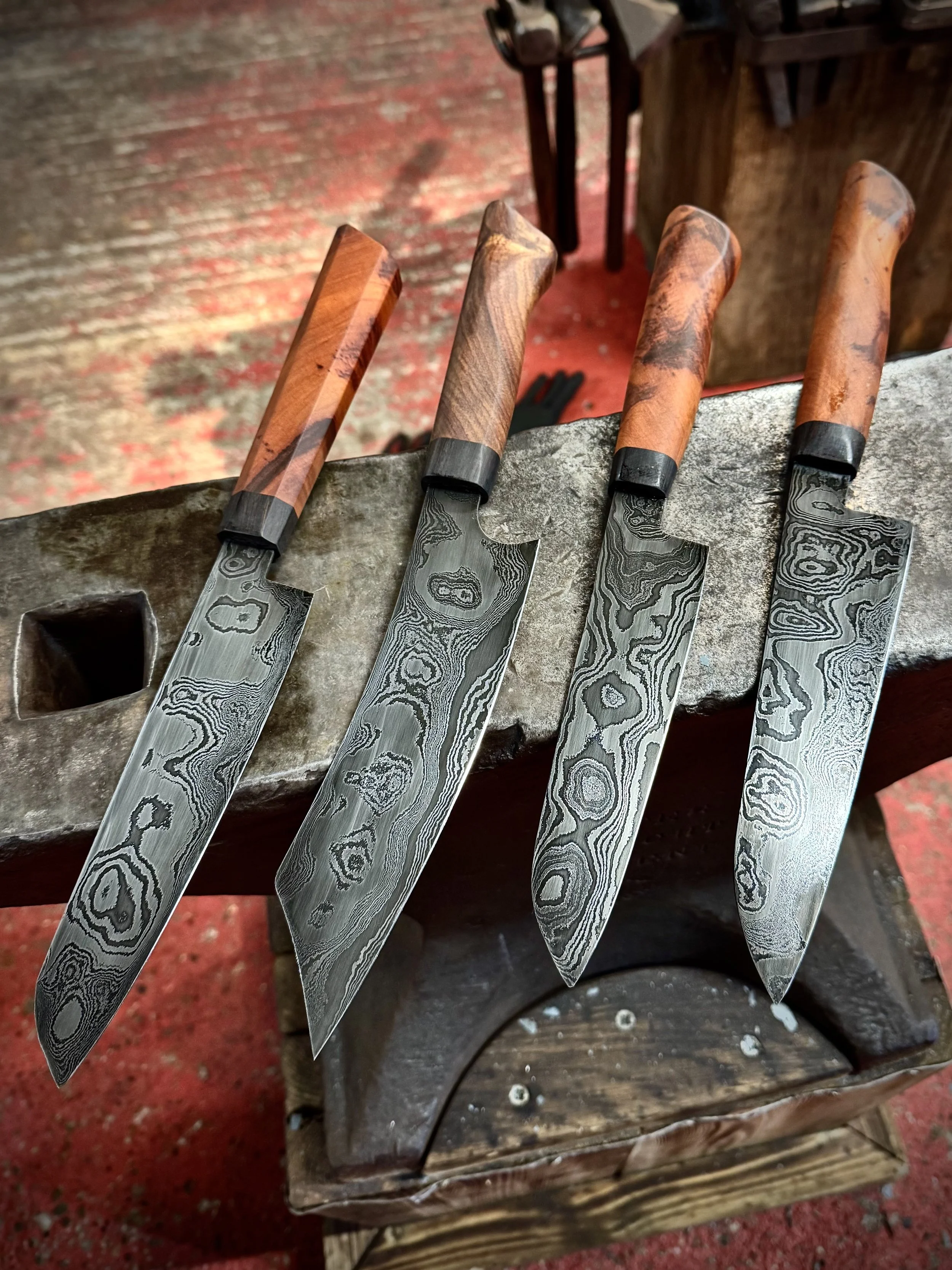 Four Damascus steel kitchen knives with wooden handles, resting on an anvil in a workshop setting.