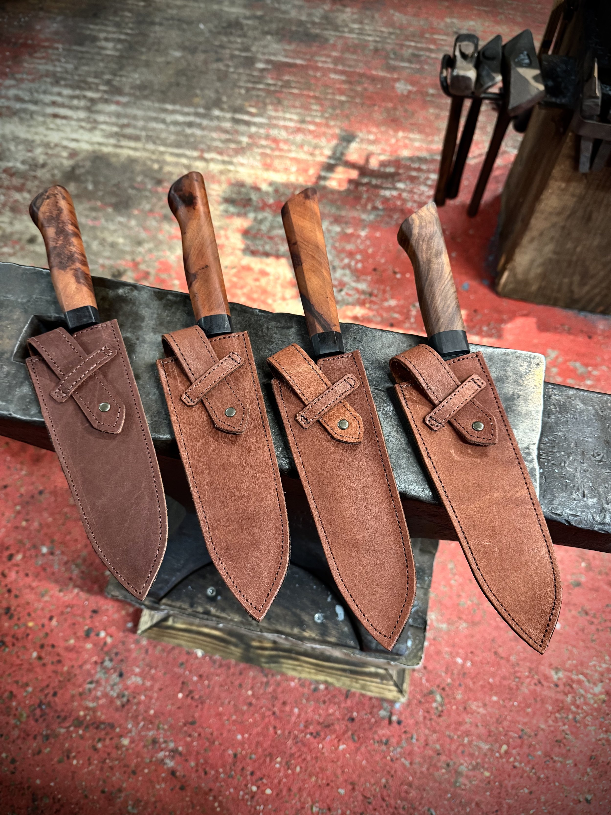 Four kitchen knives with wooden handles and leather sheaths, arranged on a workbench in a workshop setting.