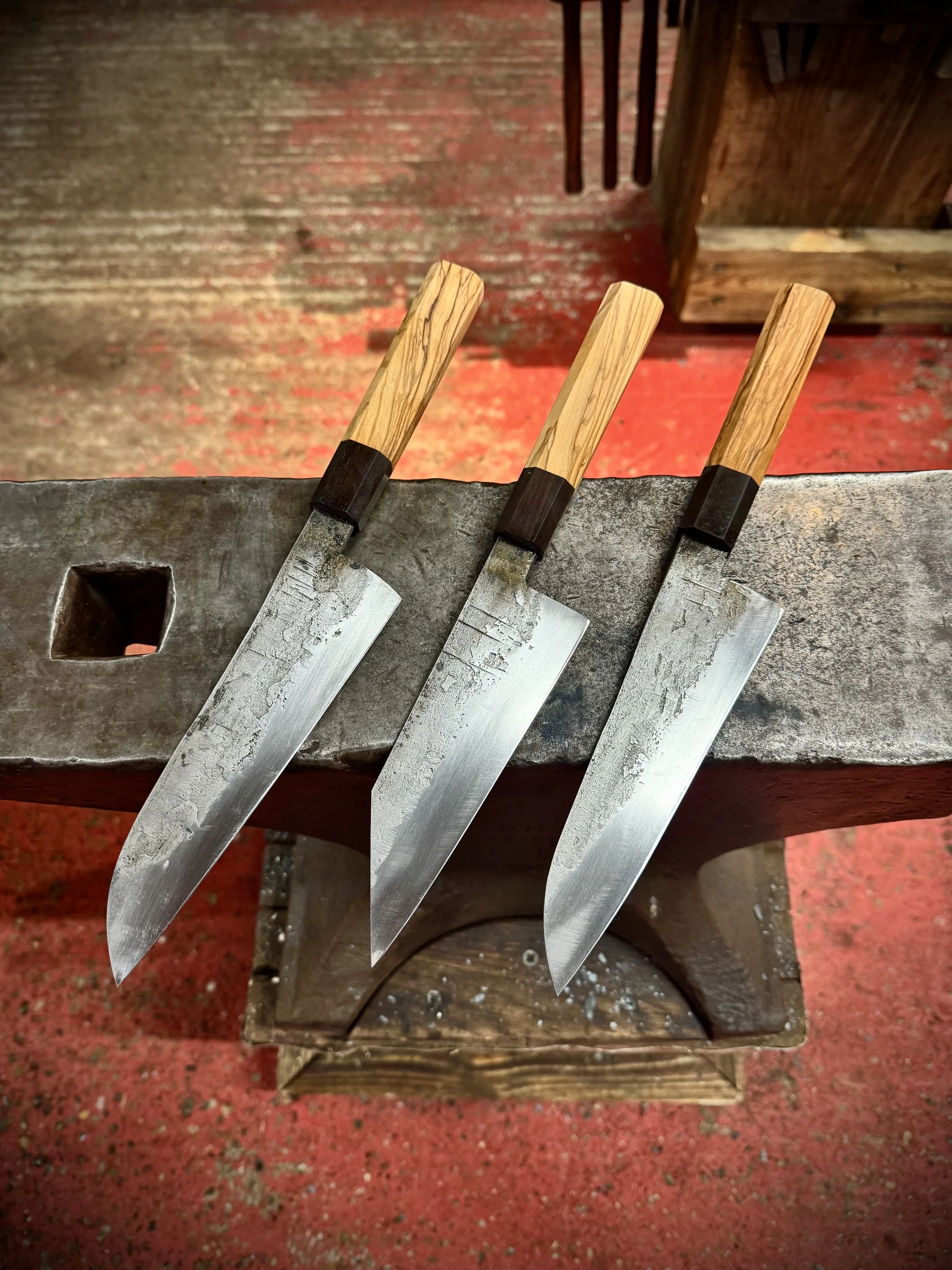 Two stainless san-mai knives with wooden handles resting on an anvil in a blacksmith's workshop.