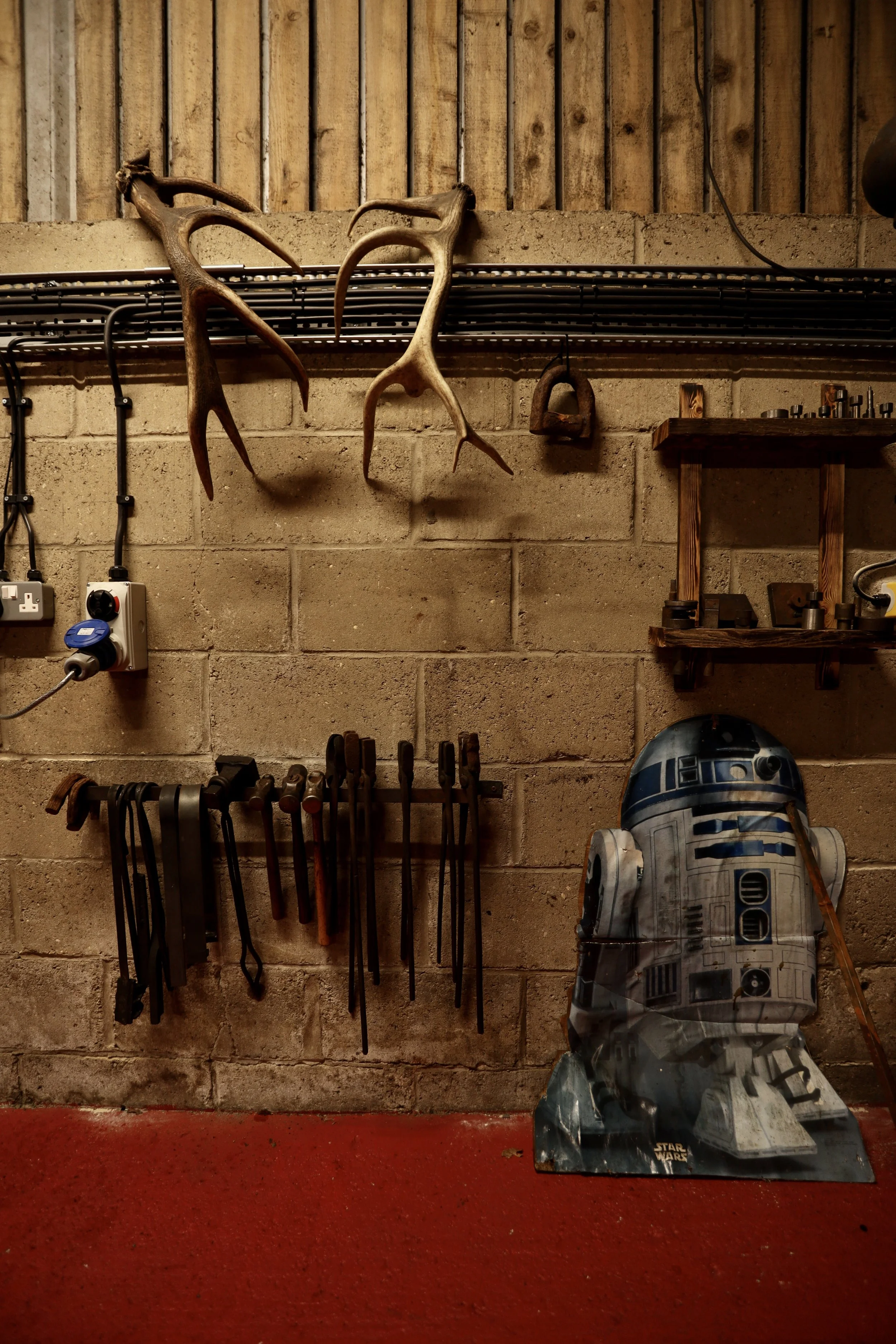 Tools and objects in a workshop including antlers hanging on the wall, a R2-D2 poster on the floor, and various hand tools hanging on a wall and a wooden shelf.