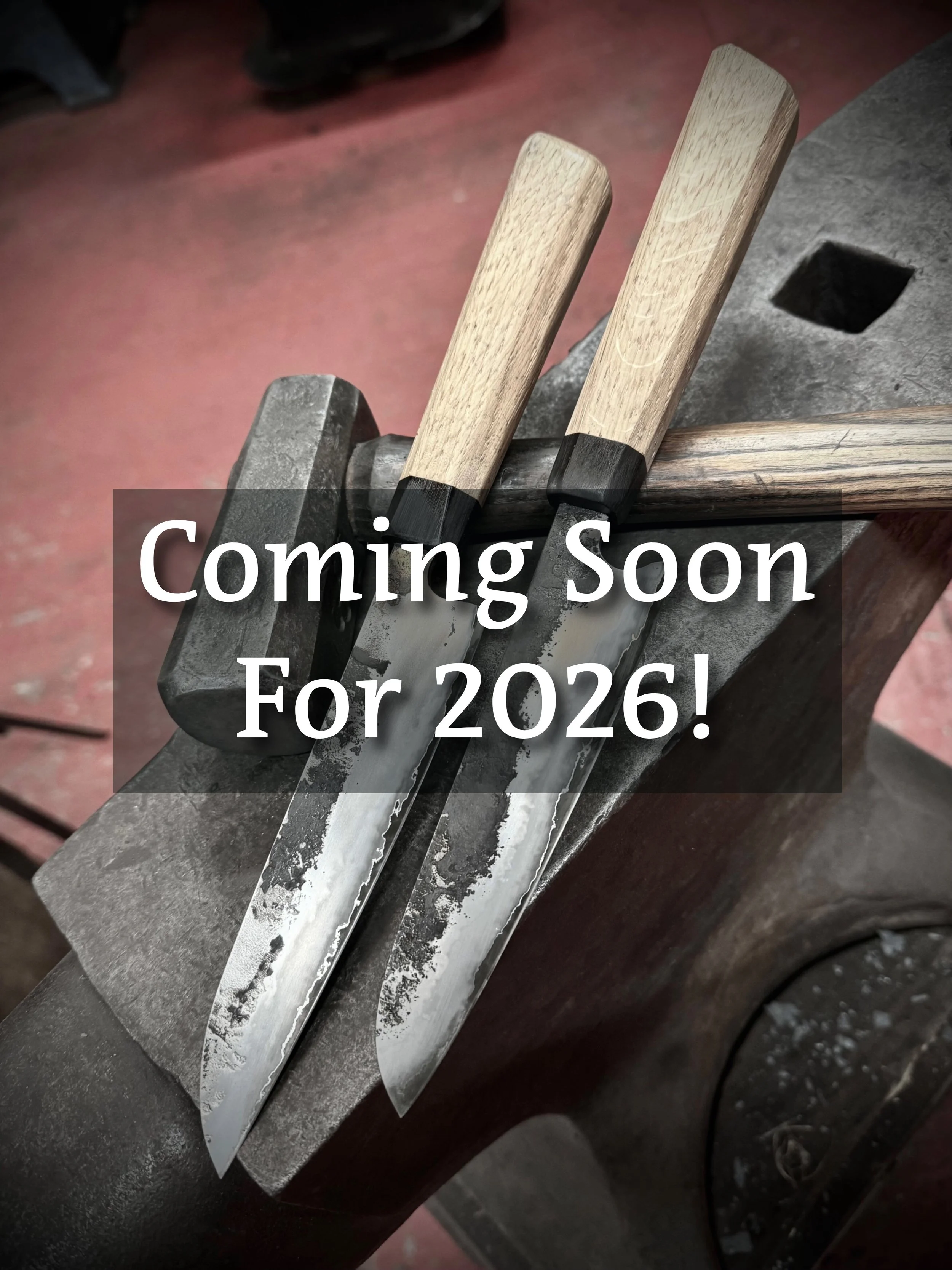 Two stainless san-mai knives with wooden handles resting on an anvil in a blacksmith's workshop.