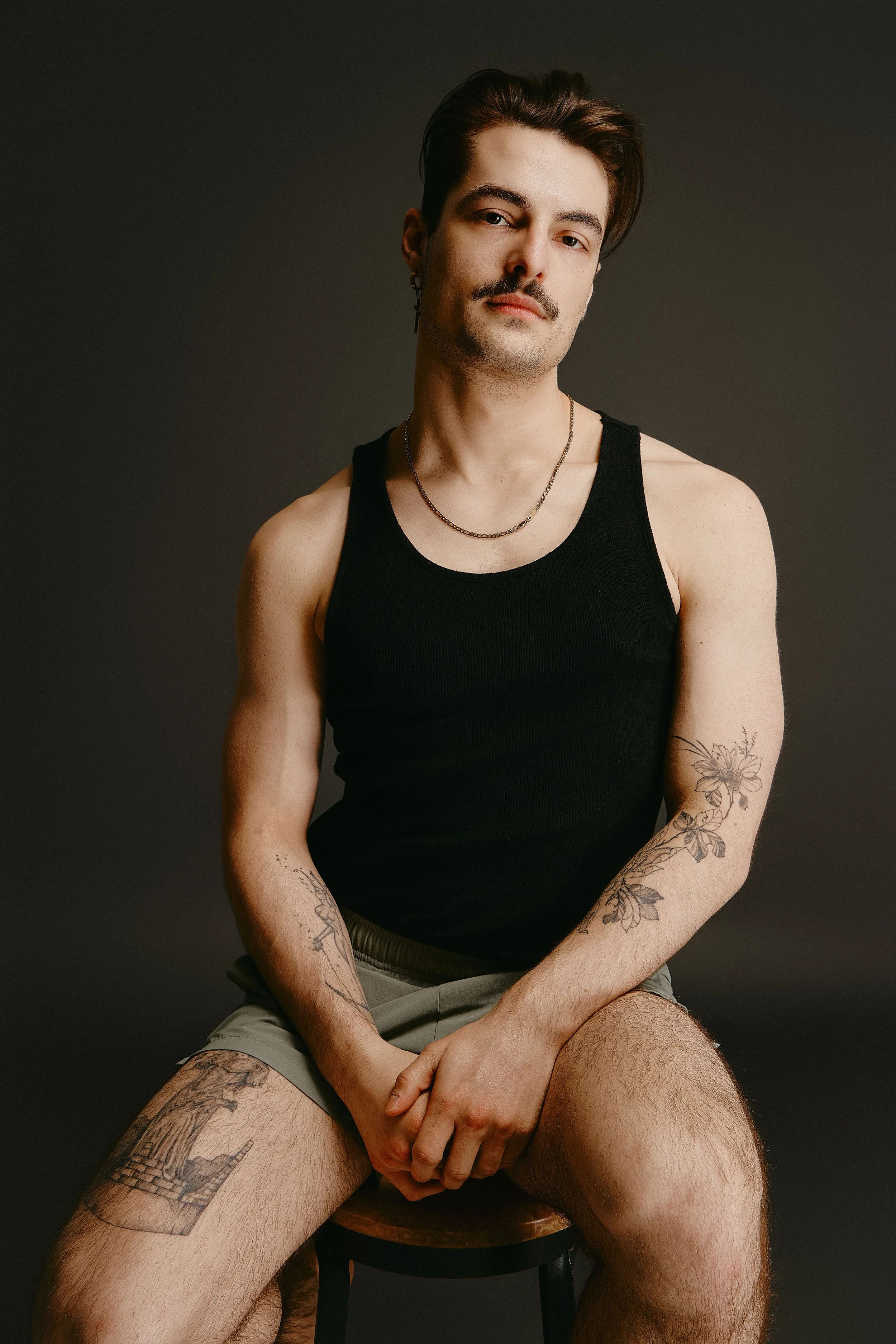 A young man with dark brown hair, a mustache, and tattoos on his arms and legs, sitting on a stool against a dark background. He is wearing a black tank top, khaki shorts, necklace, and earrings, with a confident expression.