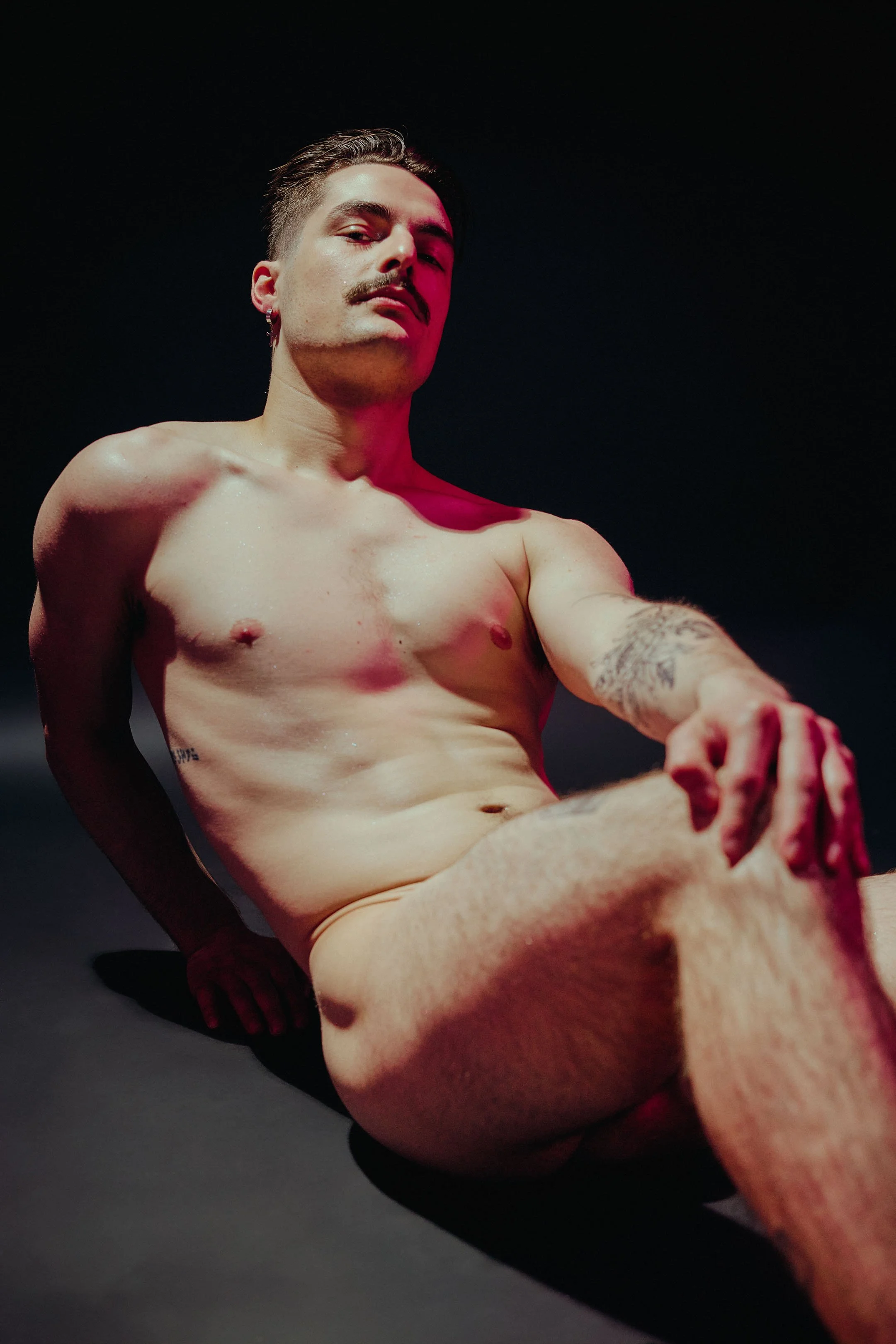 A shirtless man with a mustache and earrings sitting on a black surface, with a dark background and pinkish lighting on his face and body.