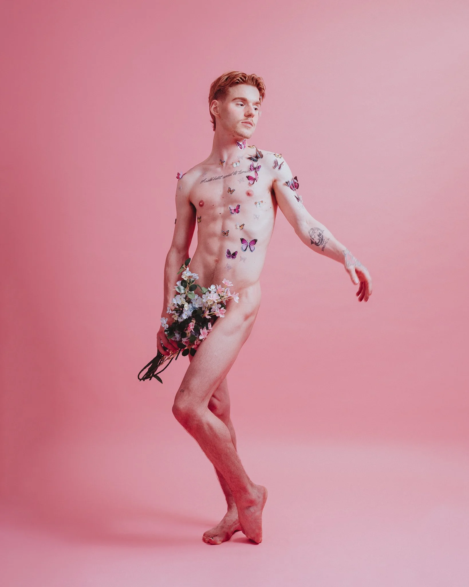 A shirtless man with short red hair stands against a pink background, adorned with colorful butterflies and flowers on his body.