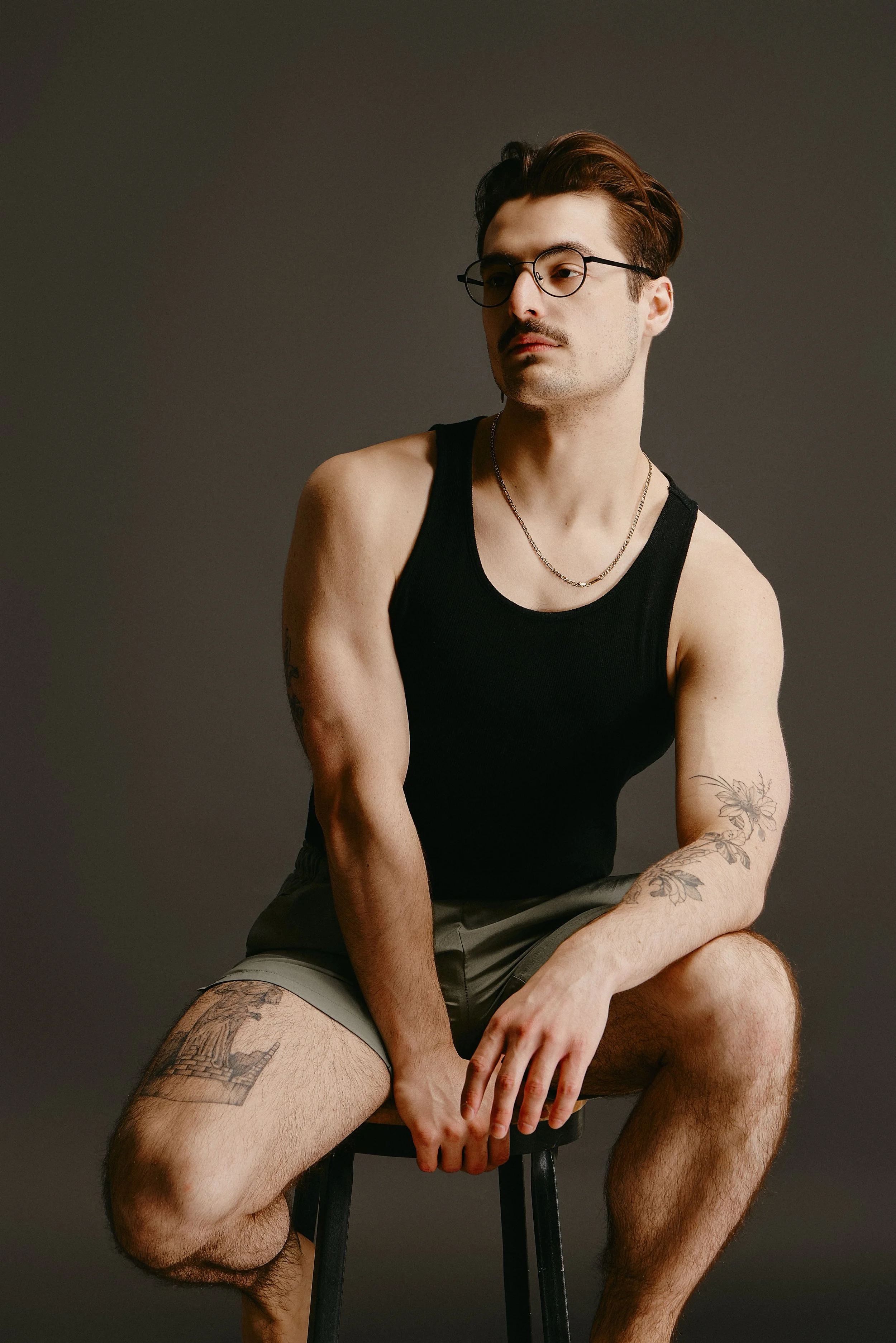 A young man with tattoos and glasses wearing a black tank top and shorts, sitting on a stool against a gray background.
