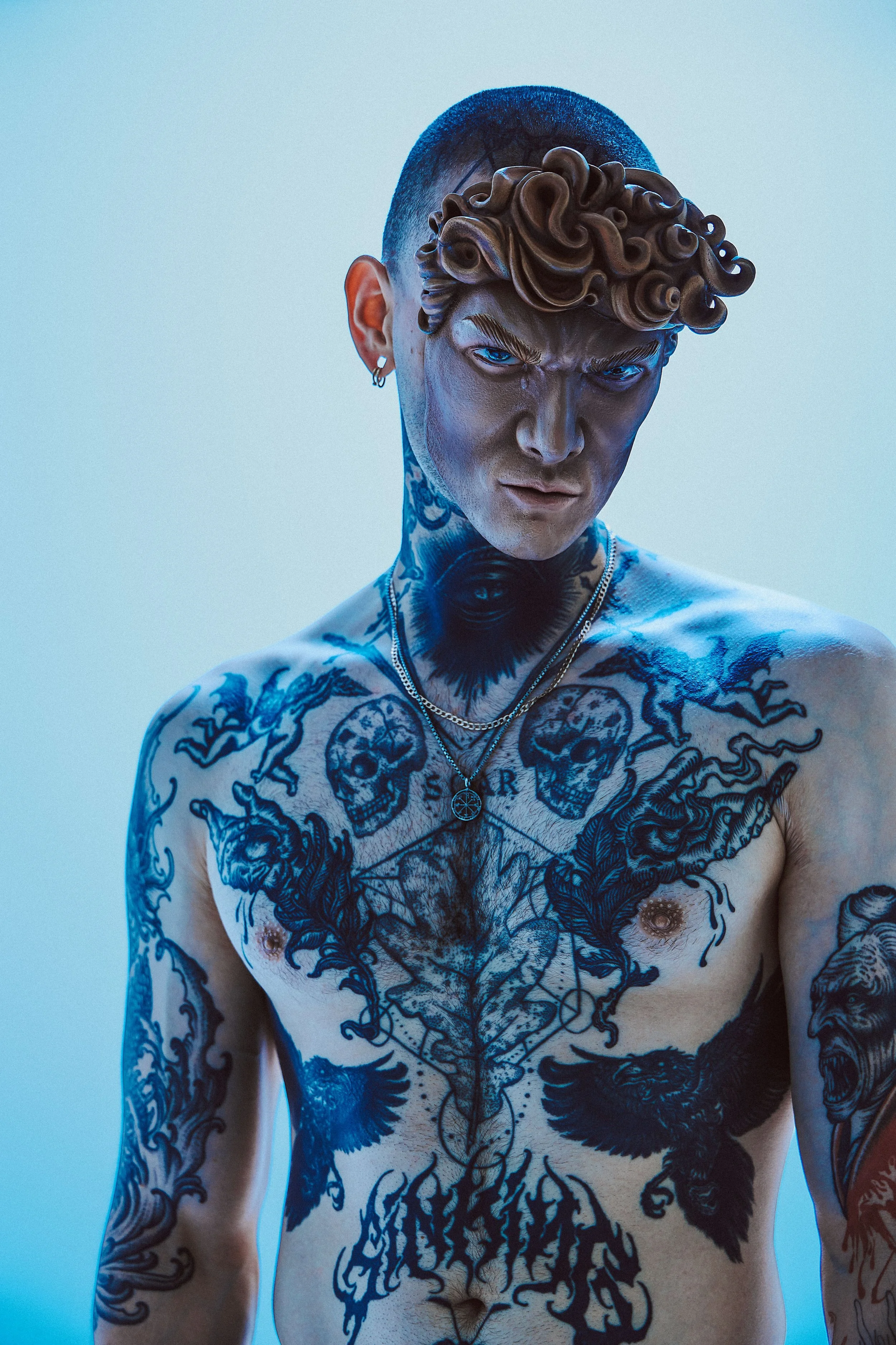 A shirtless man with tattoos covering his upper body and neck. He has short, curly hair on top and shaved sides, and is wearing jewelry including earrings and necklaces. The background is a plain light blue color.