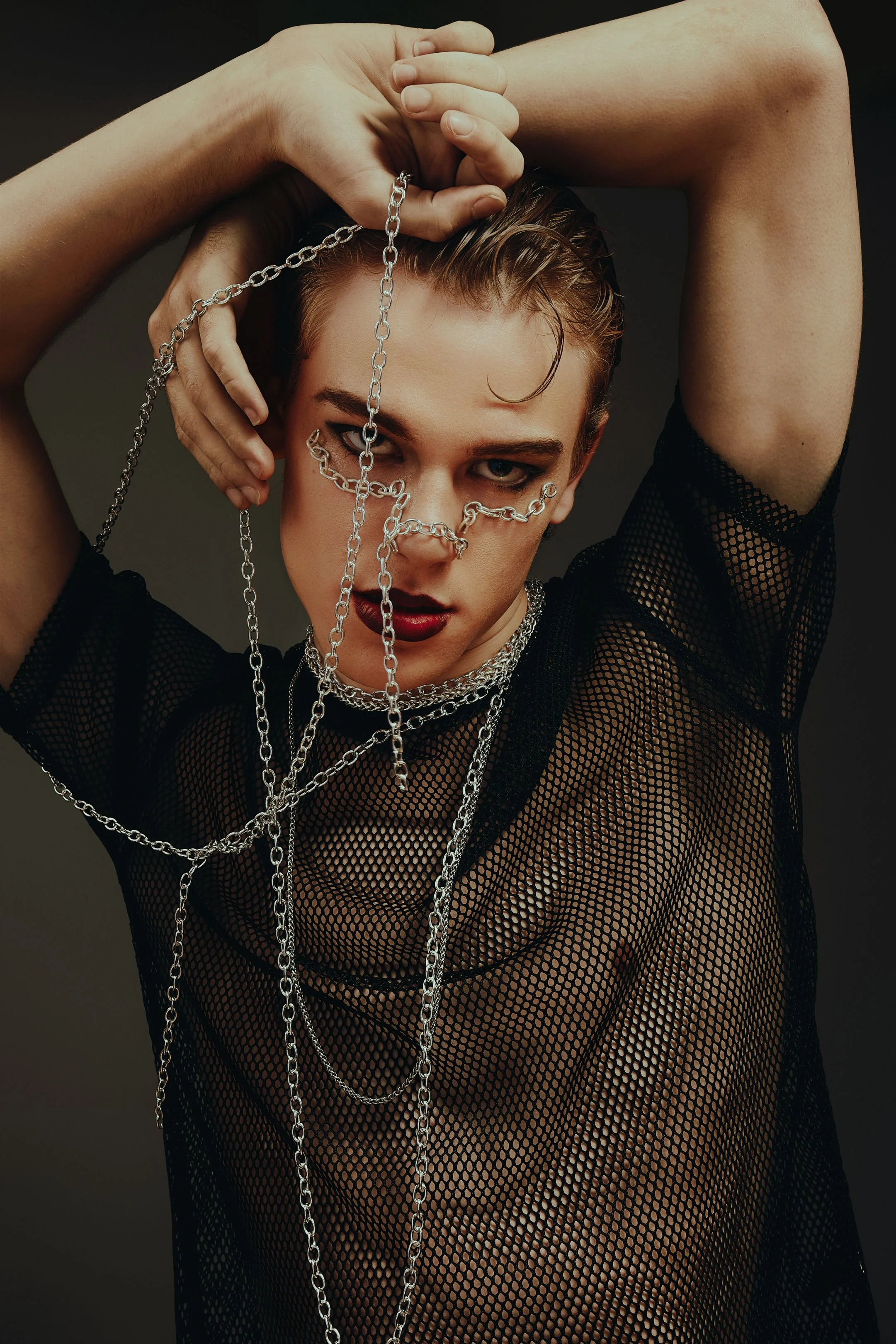 A person with short wet hair wearing dark makeup, a black mesh top, and layered silver chains, posing with their arms raised and holding chains in their hands.