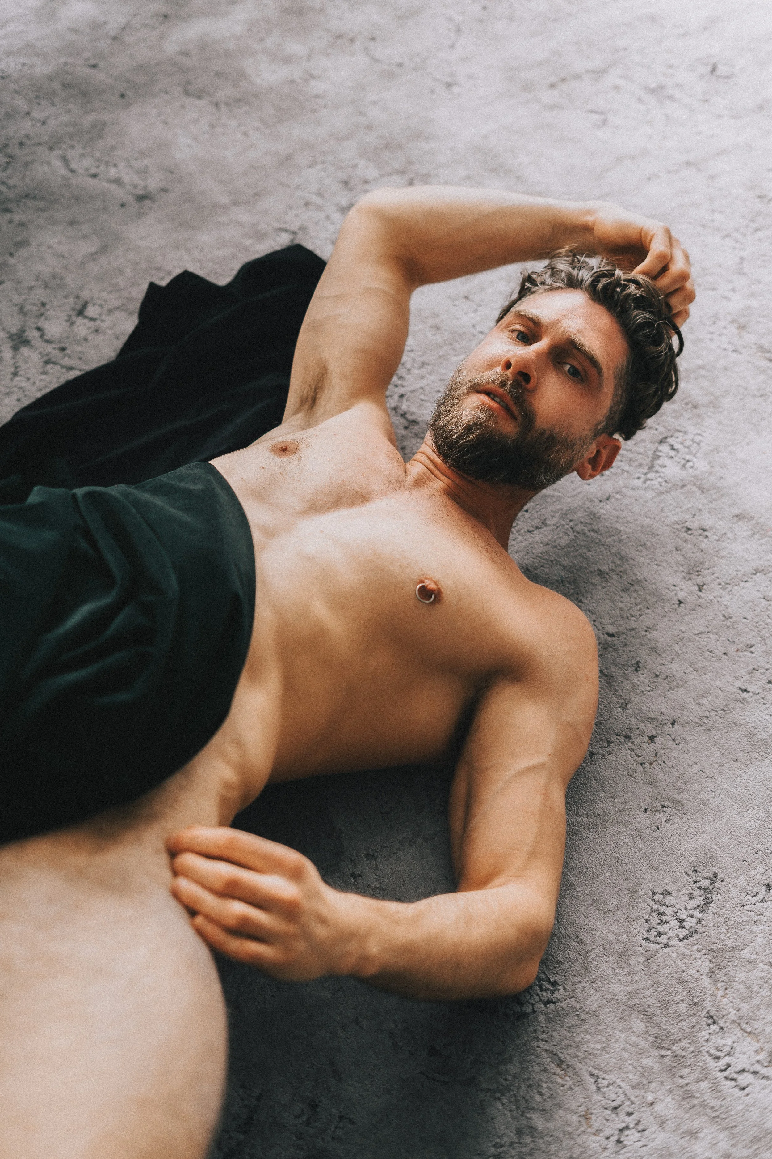 A shirtless man lying on a textured grey surface, with one hand behind his head and the other resting on his hip, looking at the camera.