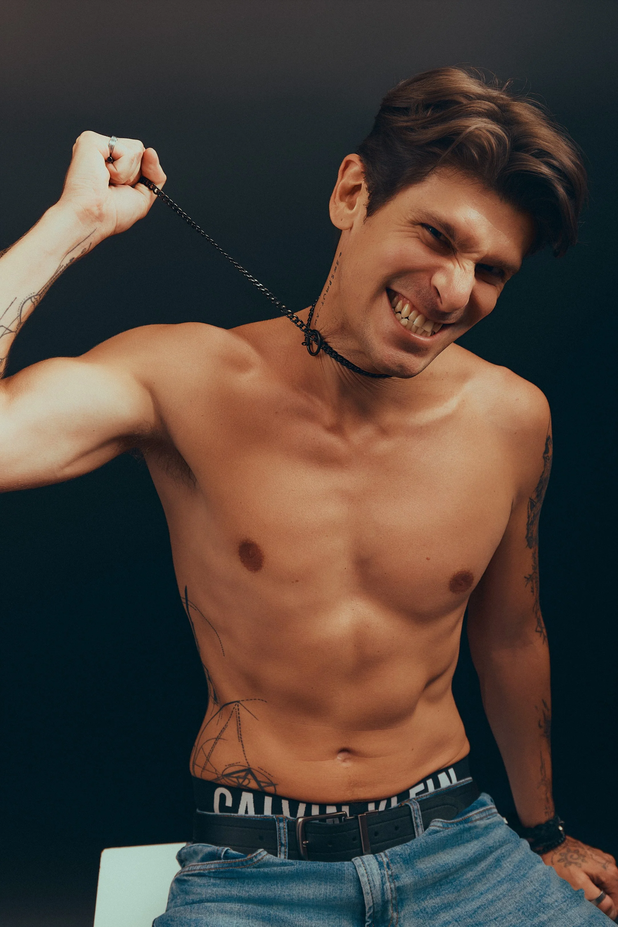A shirtless young man with tattoos, smiling and holding a black chain around his neck against a dark background.