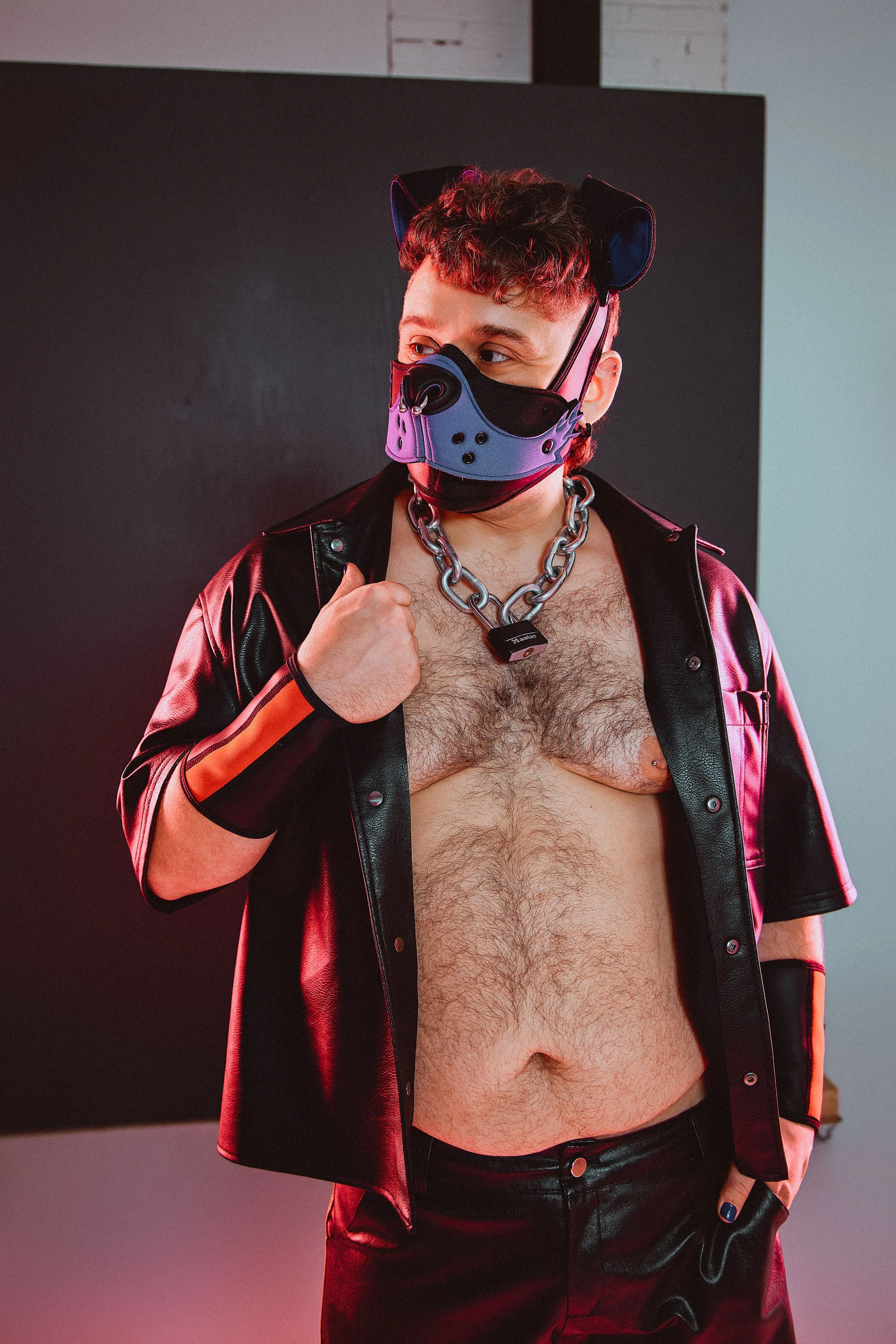 A shirtless man with chest hair wears a leather jacket, a large chain necklace, a futuristic face mask, and cat ears headband, standing against a dark background.