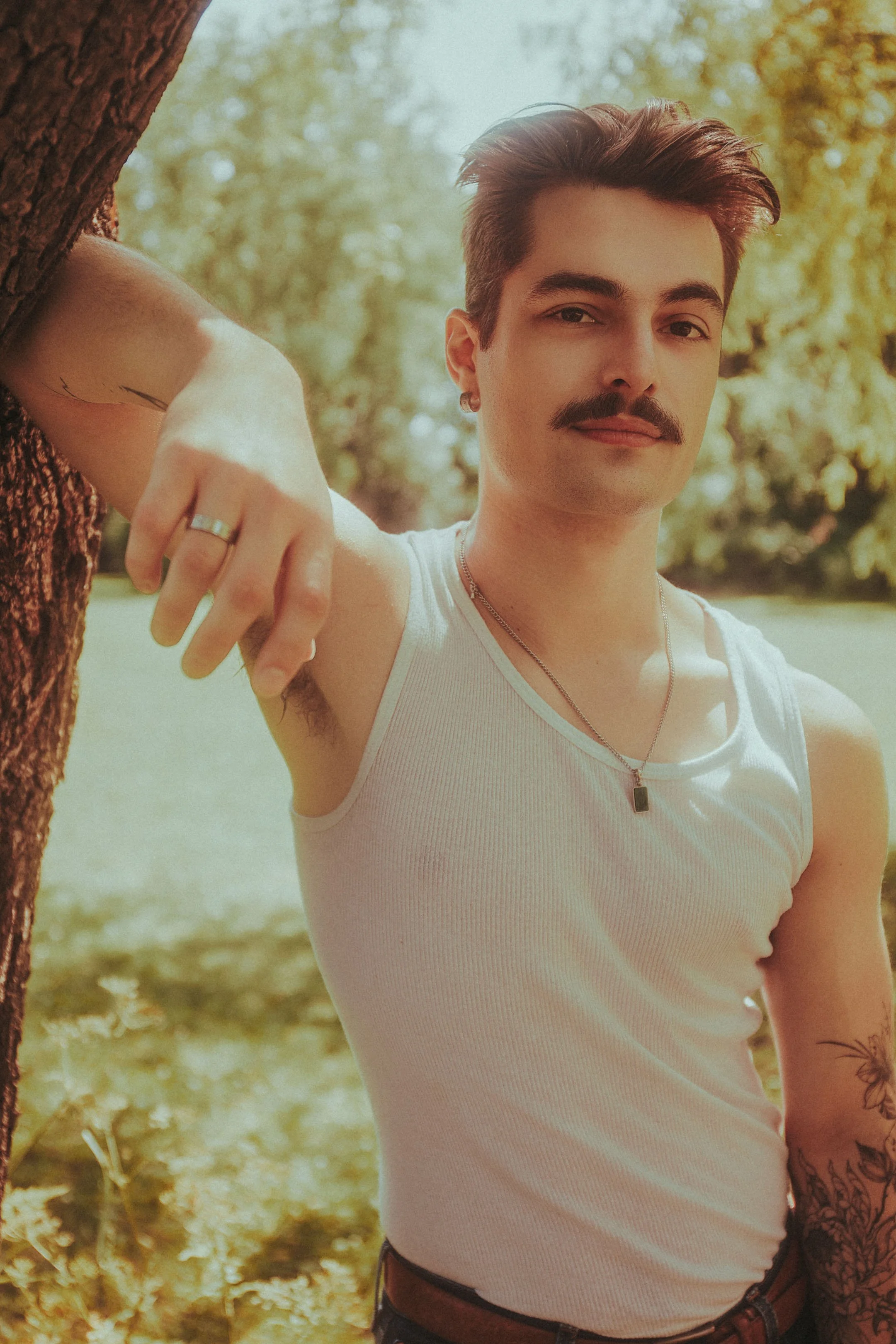 A young man with styled hair, mustache, and tattoos, leaning against a tree outdoors, wearing a white sleeveless shirt, silver necklace, and looking at the camera.