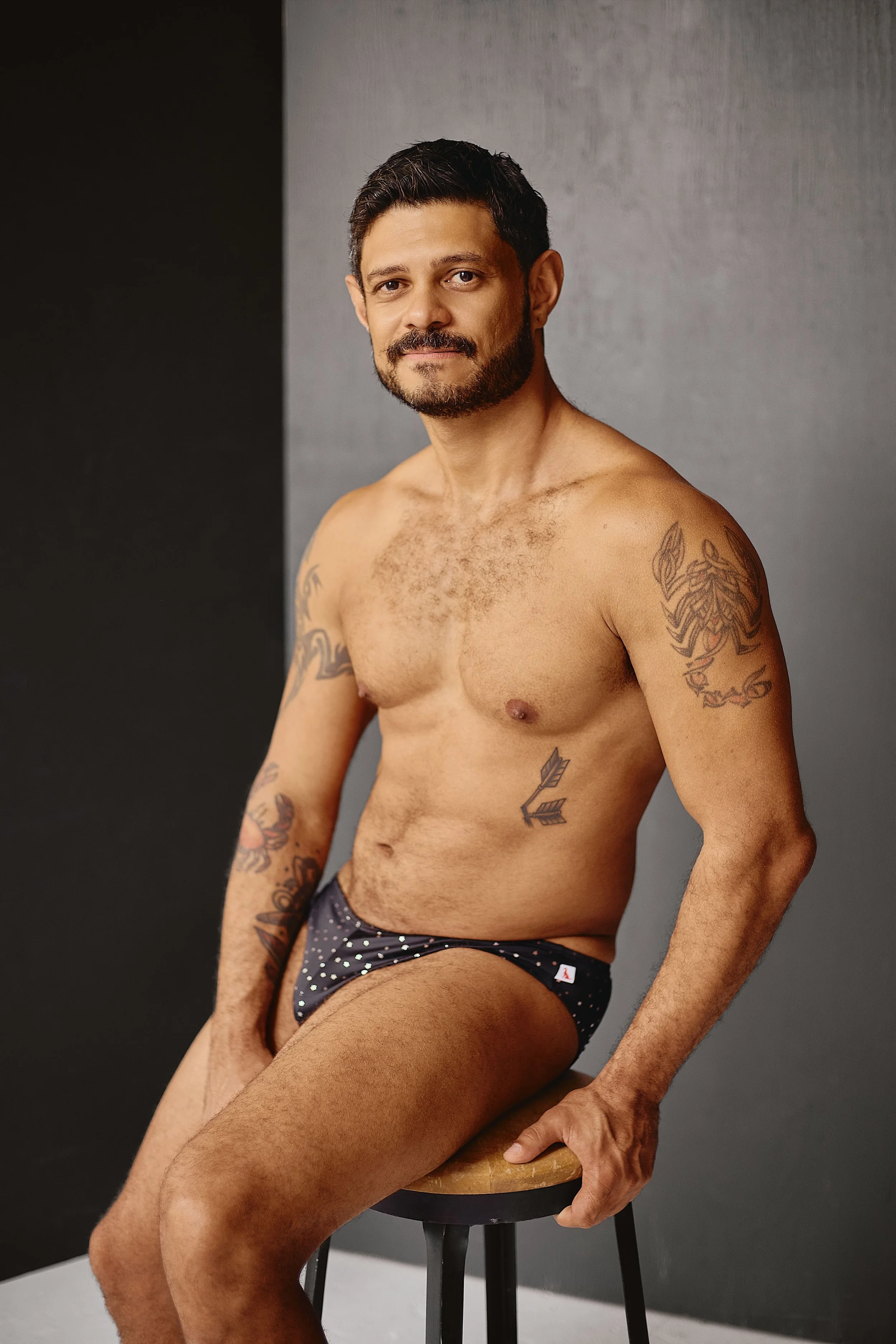 A shirtless man with tattoos on his arms and chest, wearing polka dot underwear, sitting on a stool against a gray and black background.