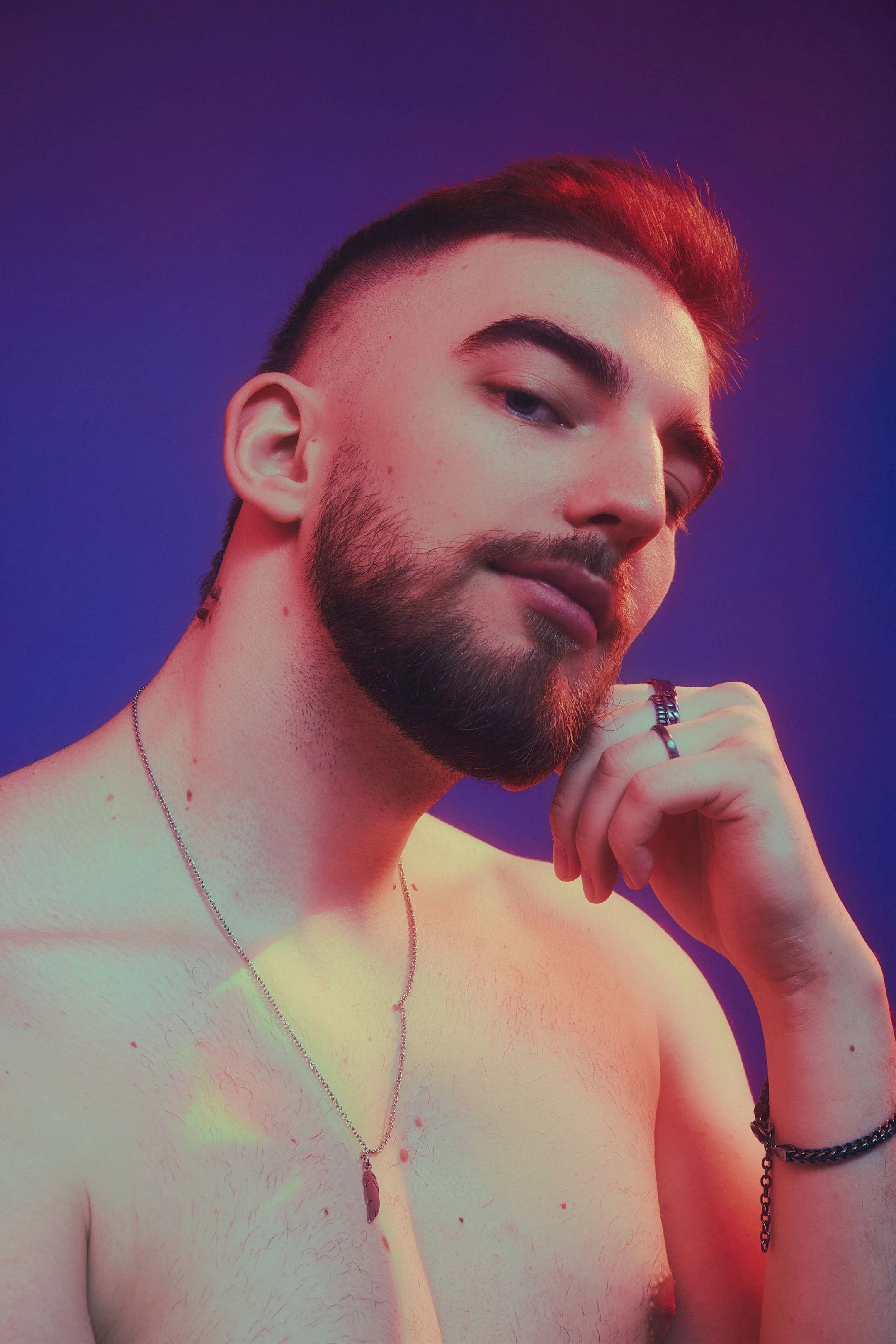 Portrait of a young man with short dark hair and a beard, posing shirtless against a purple background, wearing rings, necklaces, and bracelets.