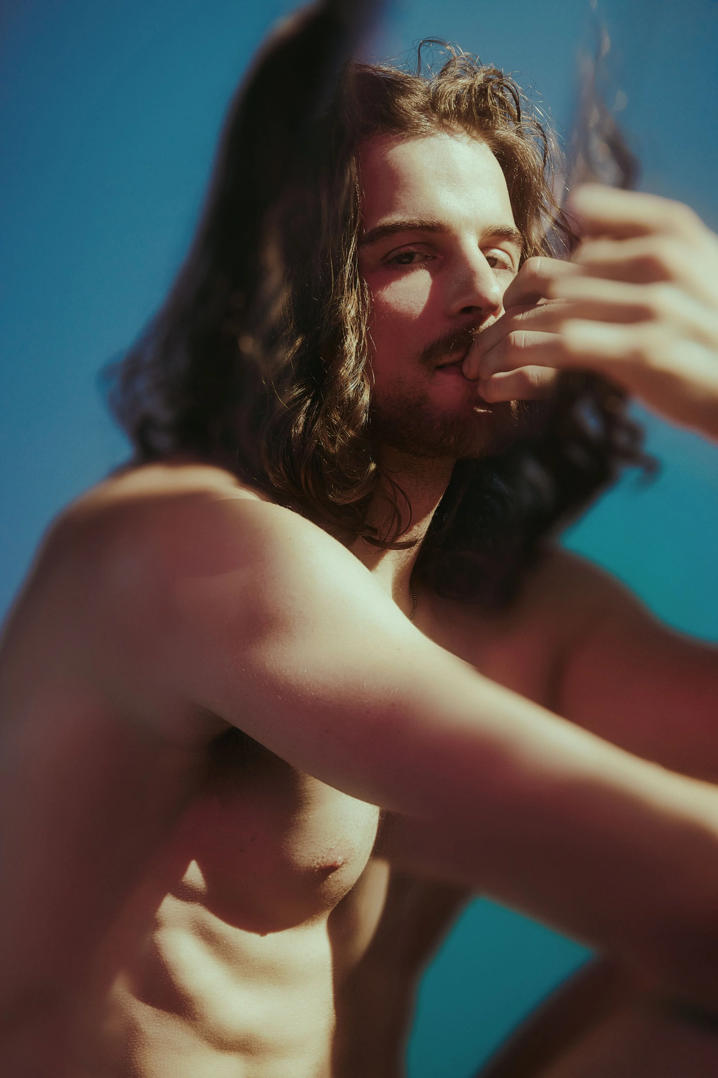 A shirtless man with long, curly hair poses against a blue sky background, touching his face with one hand.