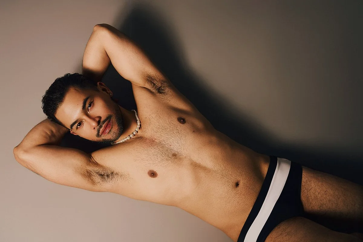 A shirtless man lying on his back with one arm behind his head, wearing a pearl necklace and dark underwear with a white waistband, against a plain background.