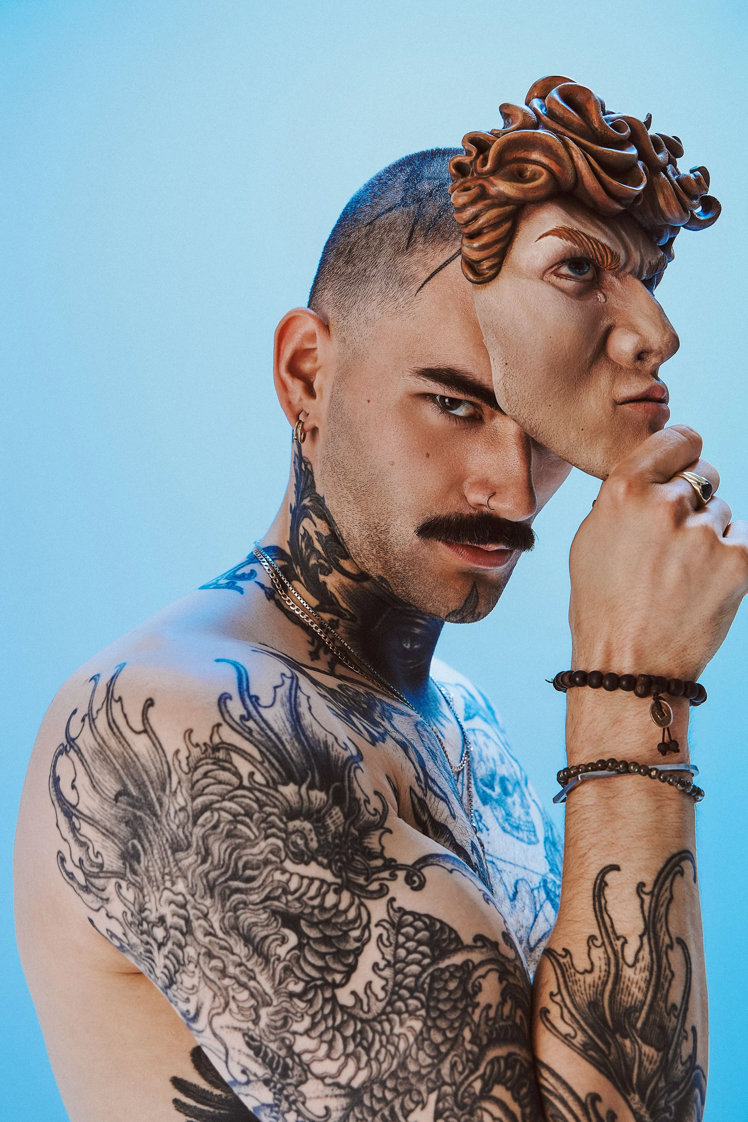 A man with tattoos on his chest and arms holds a mask of a Greek mythological figure in front of his face, against a blue background.