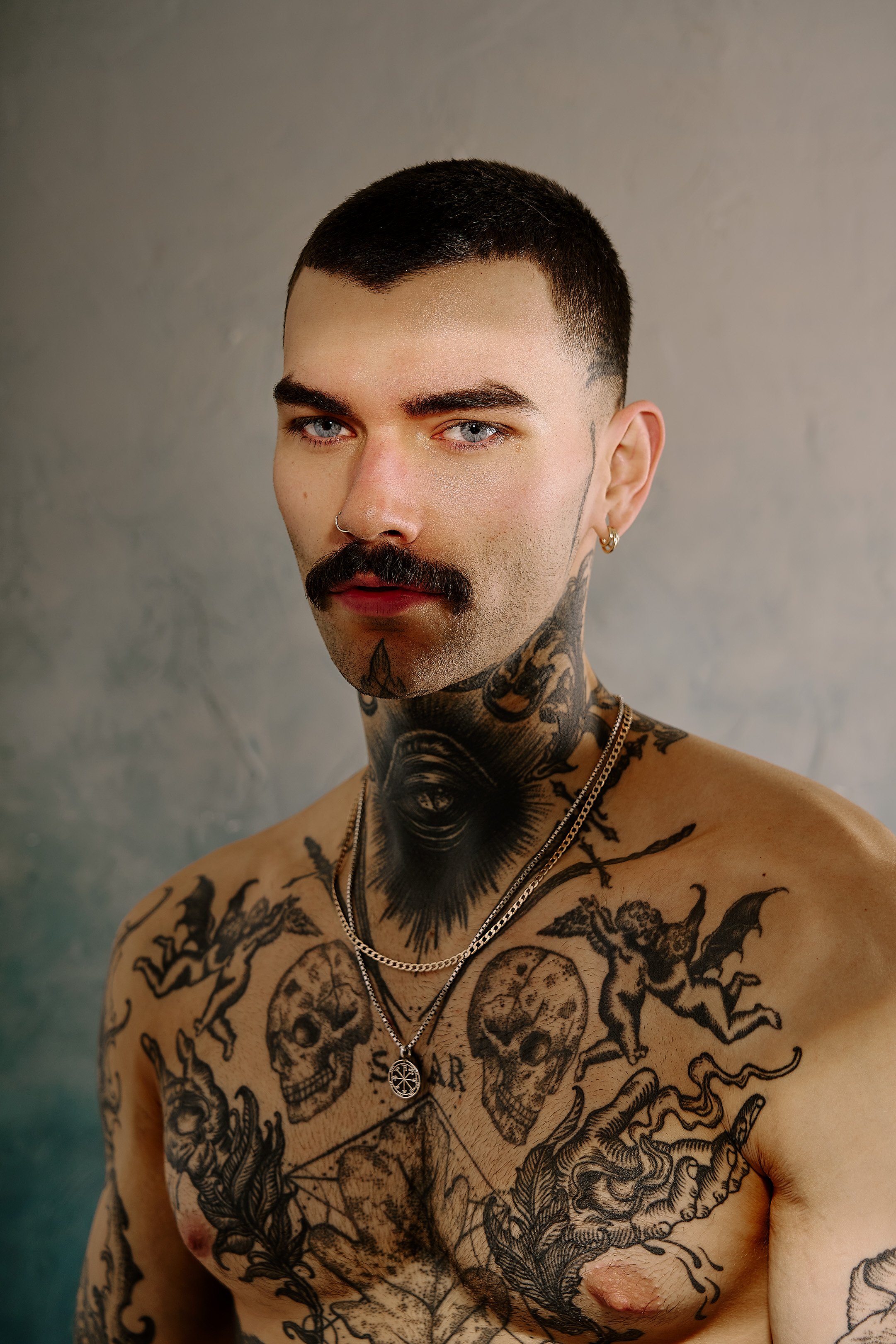 A shirtless man with tattoos on his chest and arms, dark hair, blue eyes, a mustache, a goatee, multiple earrings, and a silver chain with a pendant. The background is a plain, neutral-colored wall.