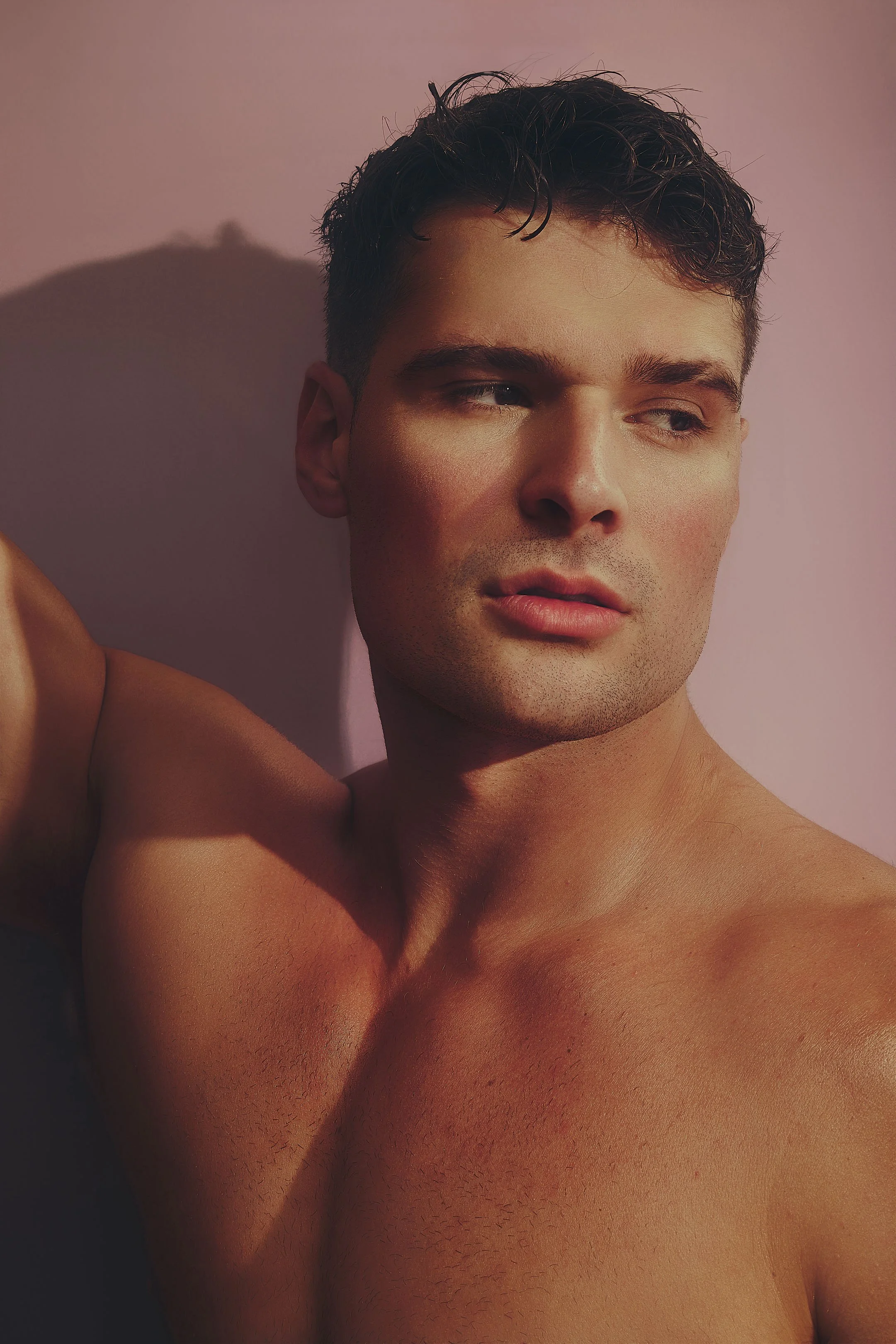 A shirtless young man with short, dark, slightly curly hair, looking to the side, against a pink wall with a shadow.