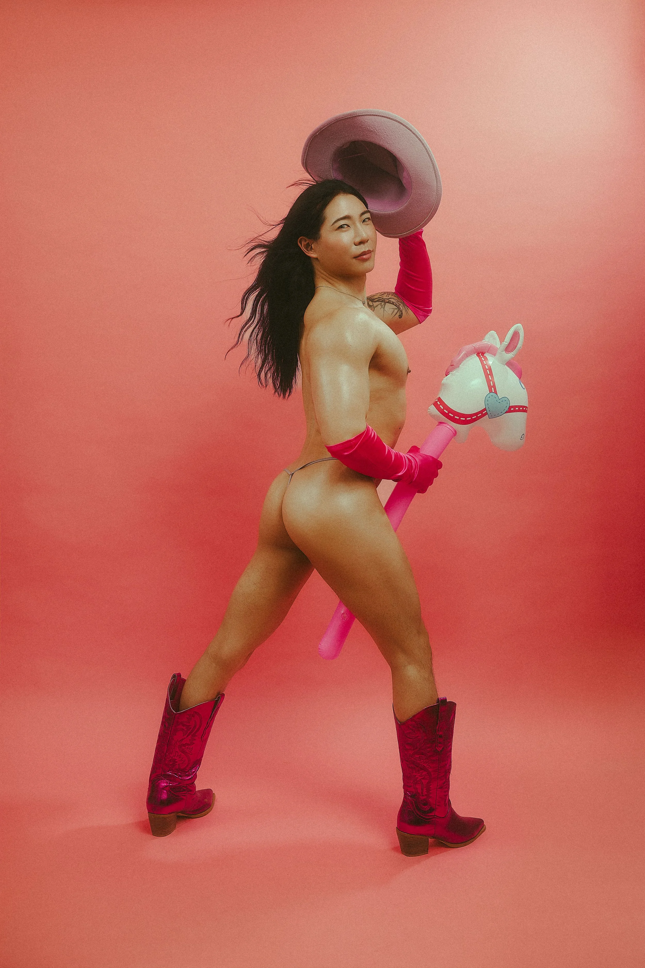 A topless woman with long black hair poses against a pink background, wearing pink cowboy boots, red gloves, and holding a pink toy horse with a heart-shaped saddle and a hat, looking at the camera.