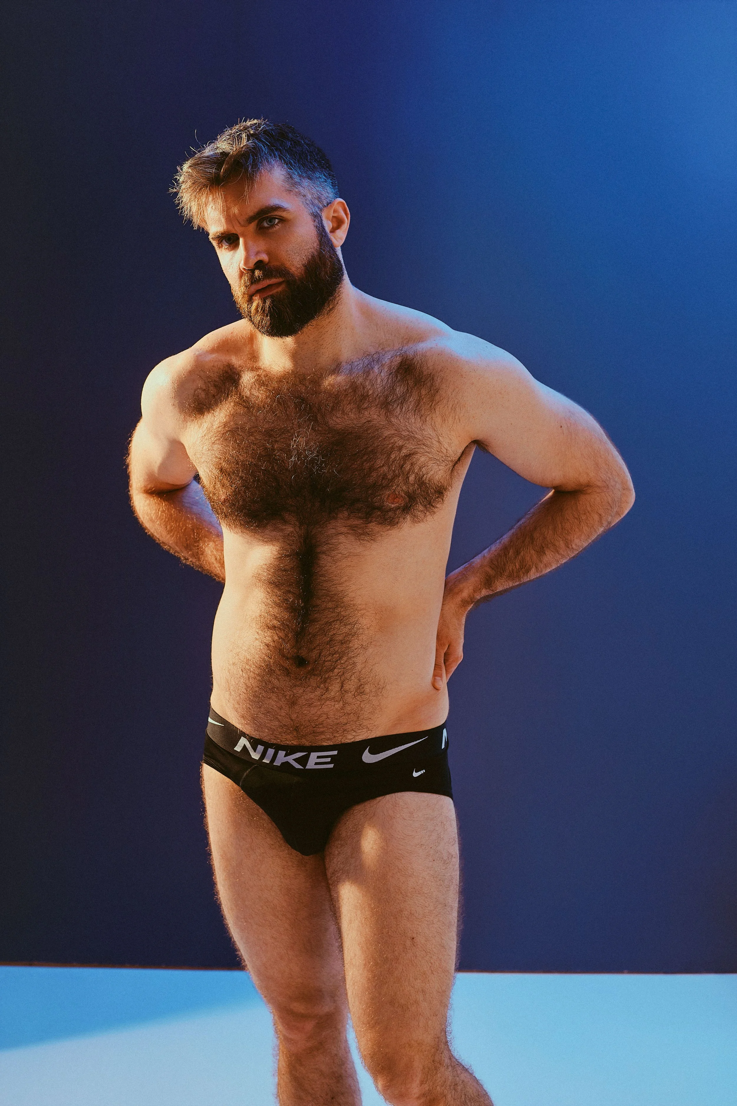 A man with a beard and dark hair standing shirtless against a blue backdrop, wearing black Nike underwear.